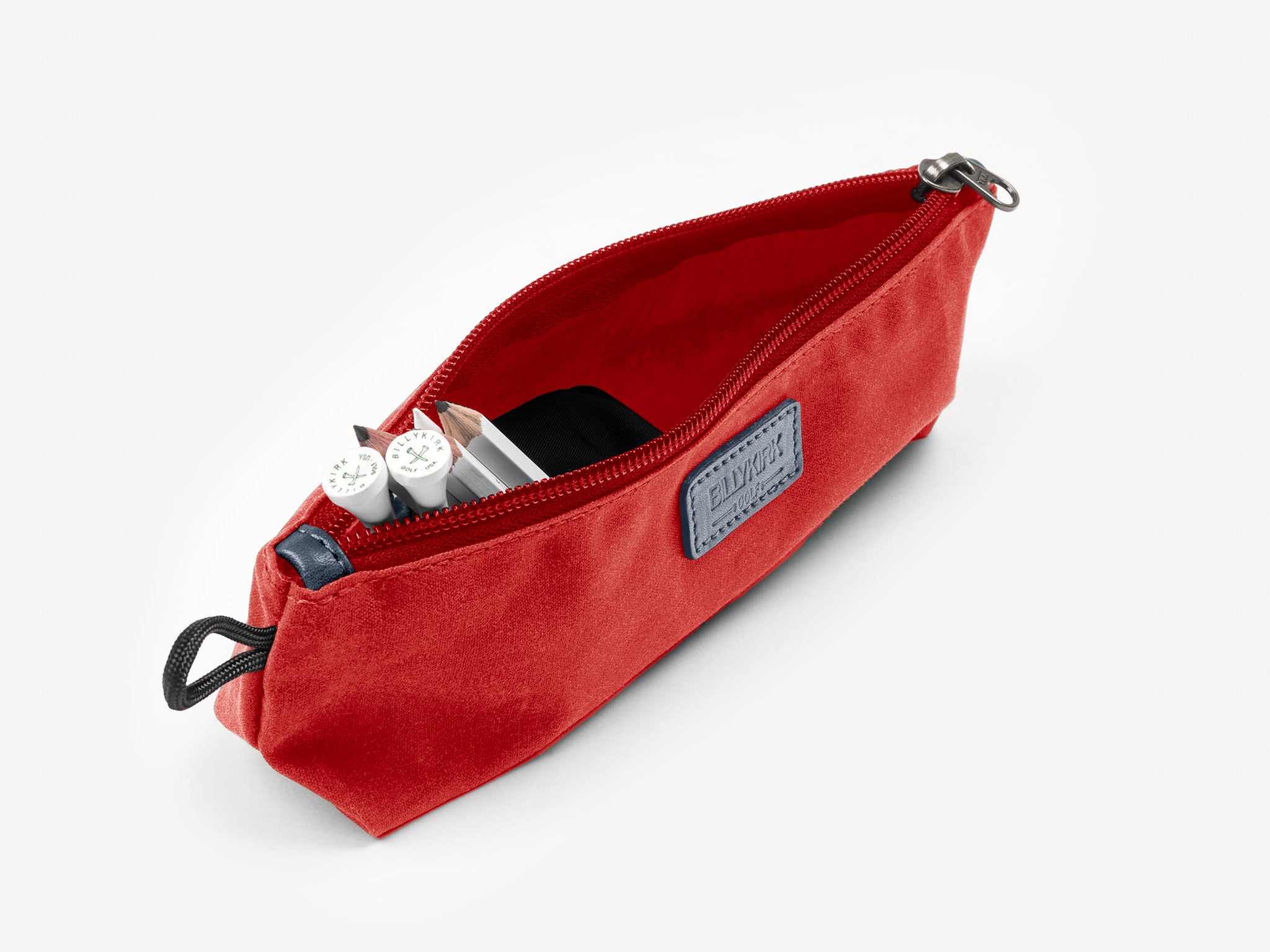 A red zippered pencil case is open, revealing white markers, a black item, and a silver pen inside. The case has a blue patch on the side and a black loop for carrying.
