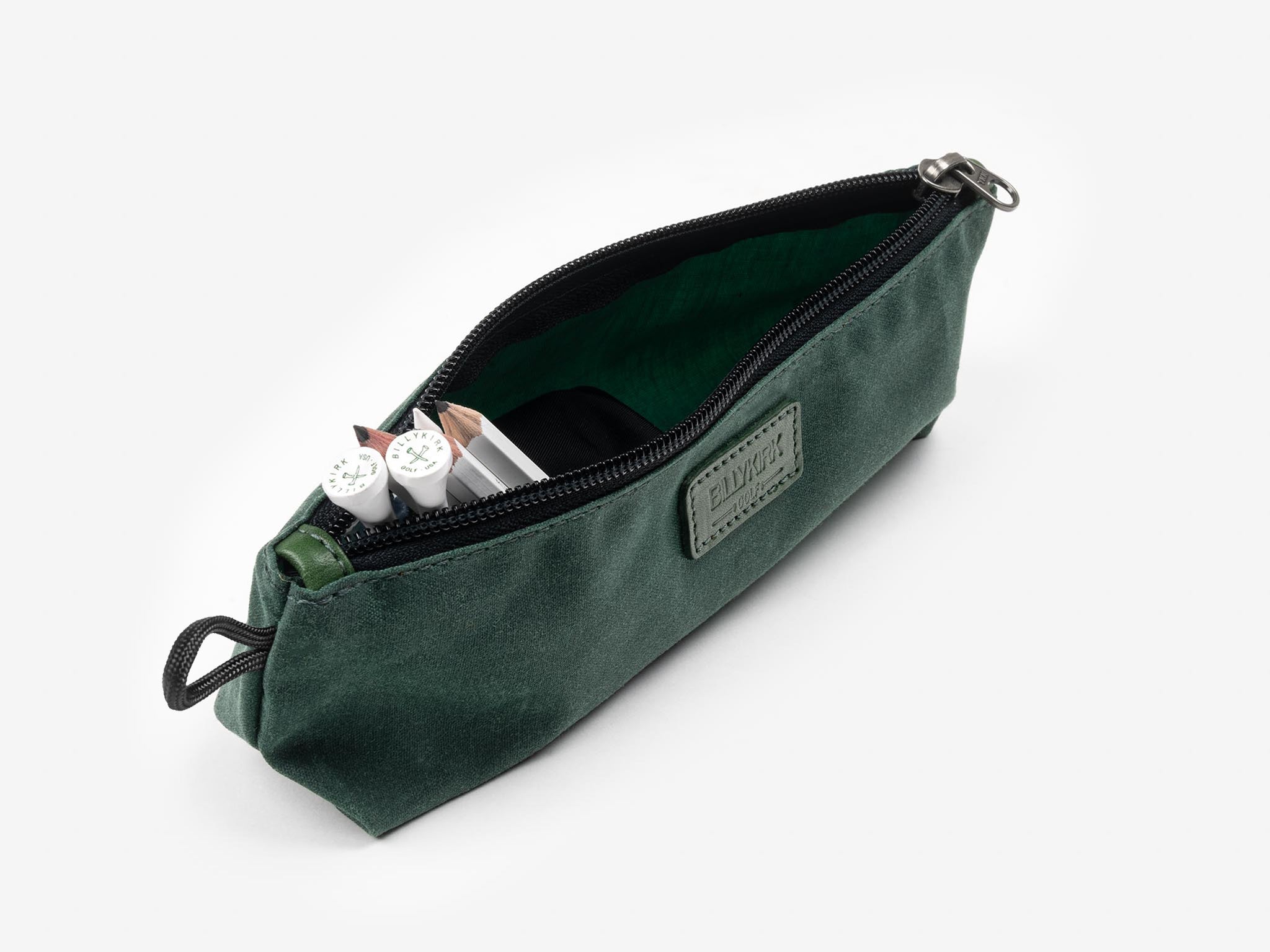 A dark green zippered pouch is open, revealing two white pens and a notebook inside. The pouch has a fabric label on the front and a loop on one end. The background is plain white.