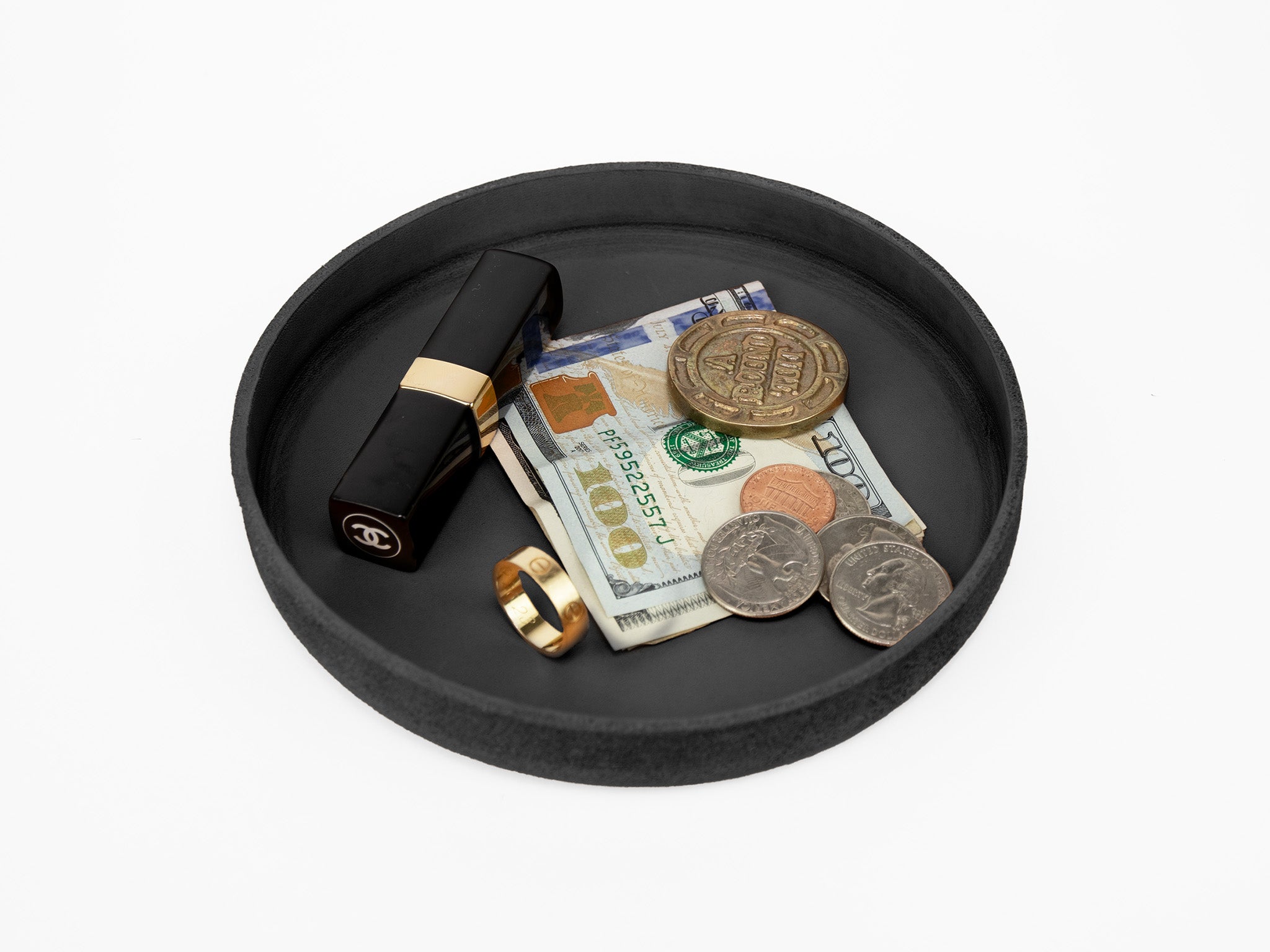 A black round tray holds a Chanel lipstick, a gold ring, a folded 100 US dollar bill, a one dollar coin, a quarter, and a ten peso Mexican coin on a white background.

