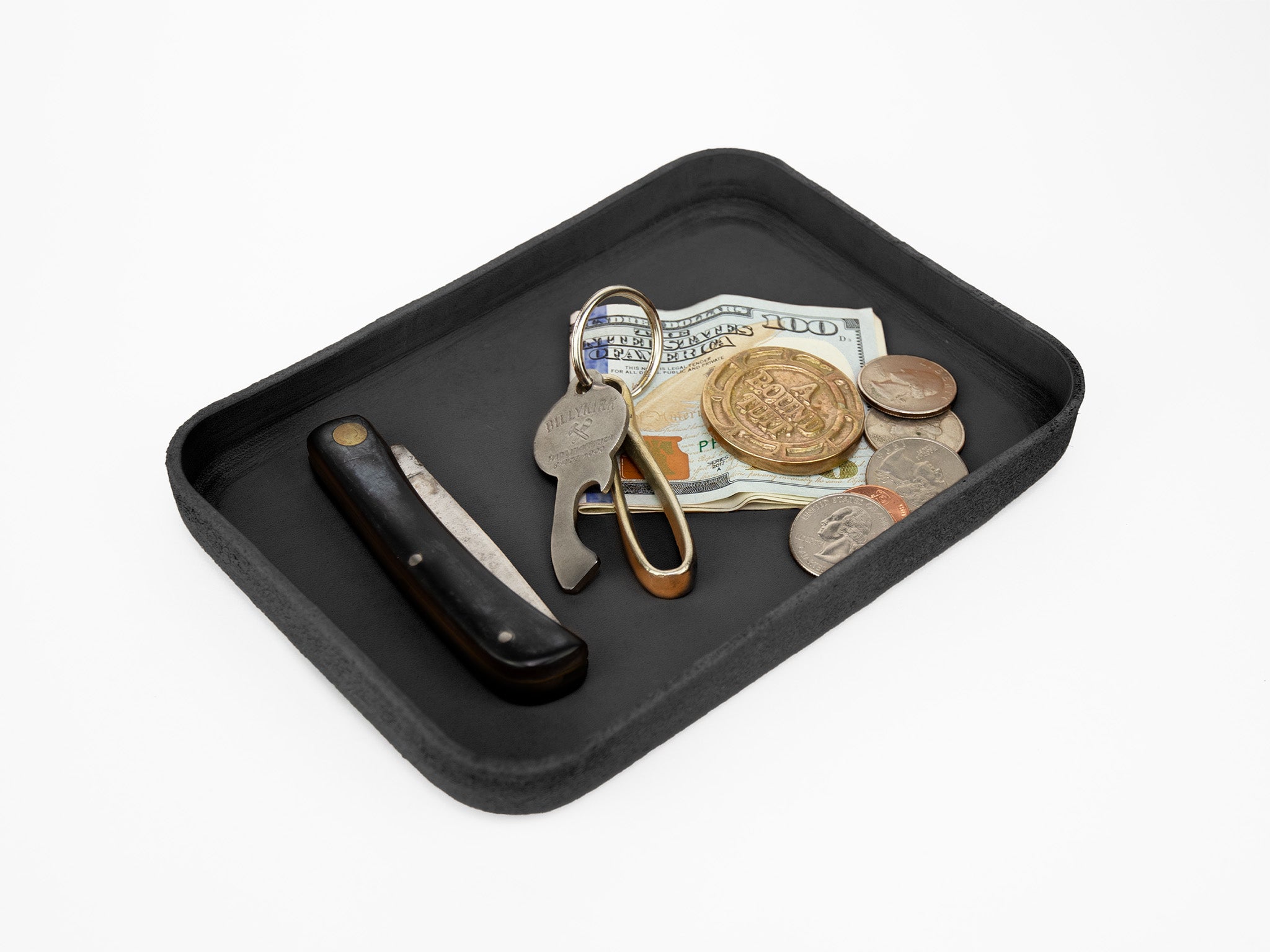 A black tray holds a folding pocketknife, a bottle opener keychain, a folded US $100 bill, coins, and other small currency on a white background.