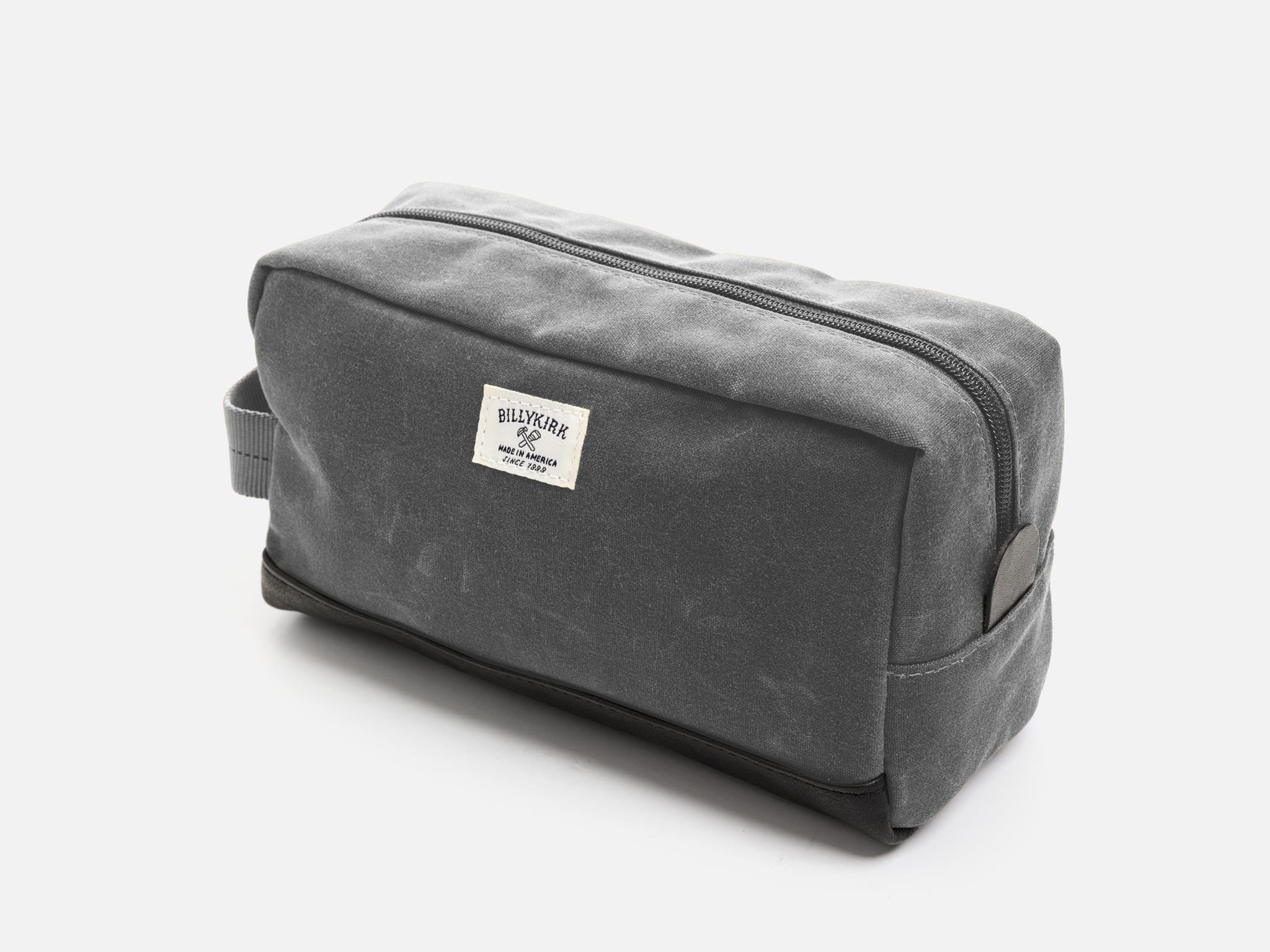 A rectangular ash grey waxed canvas toiletry bag with a zipper, a grey nylon webbing handle, a black leather base, a leather tab to help close the zipper, and an off-white Billykirk brand label on the front.