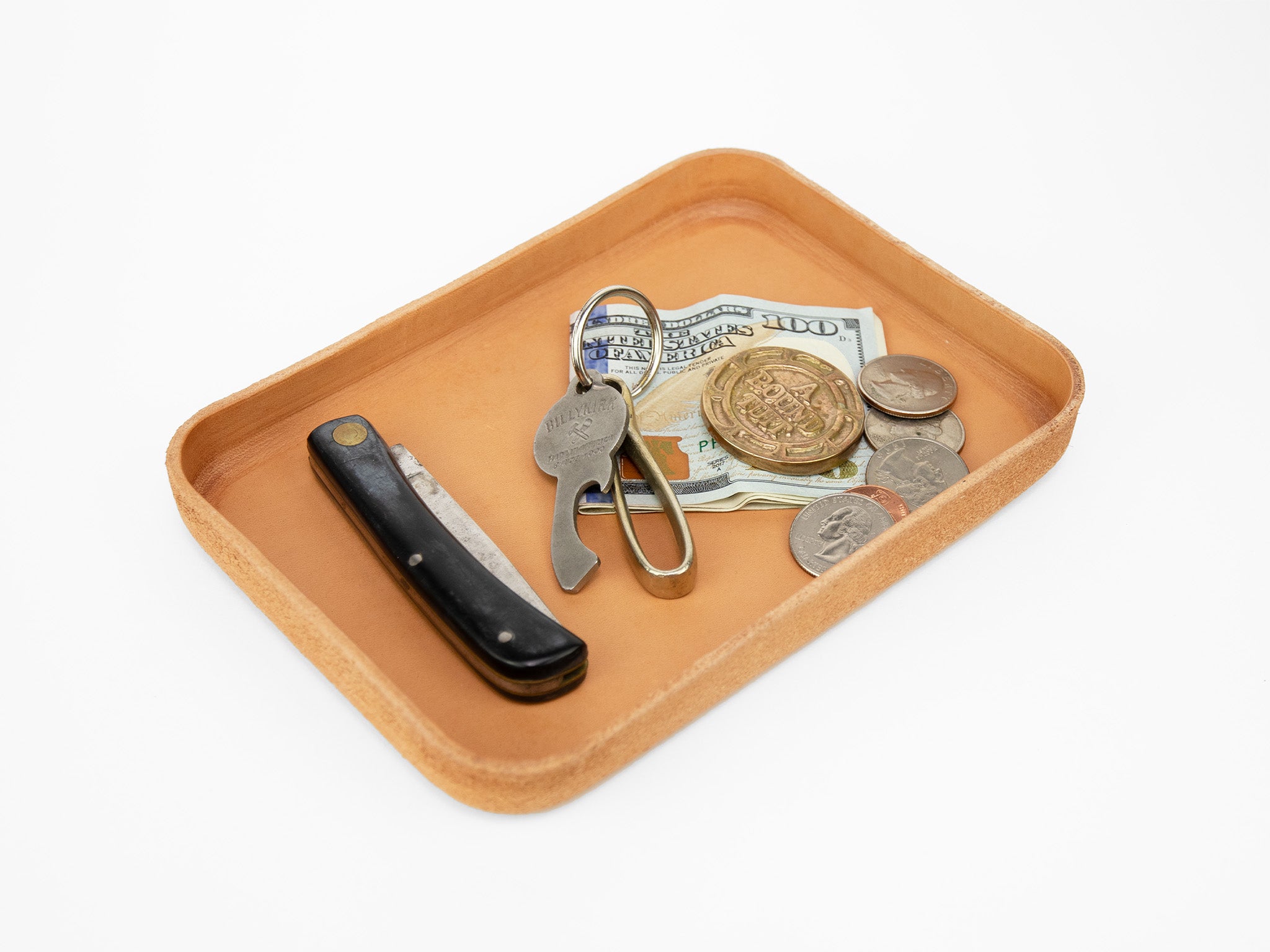 A leather tray holding a folded $100 bill, various coins, a folding knife, and a keychain with keys and a bottle opener, all placed on a white surface.