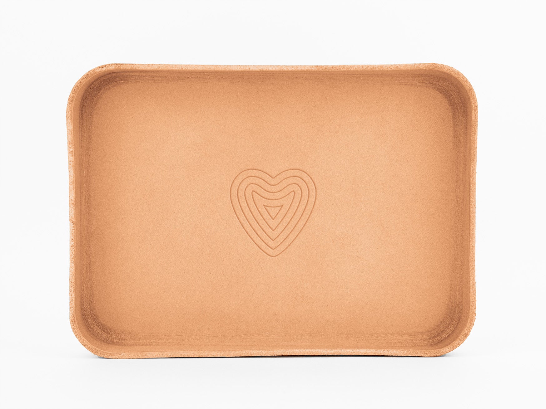 A rectangular, light brown leather tray with rounded corners, featuring an embossed layered heart shape in the center, set against a plain white background.