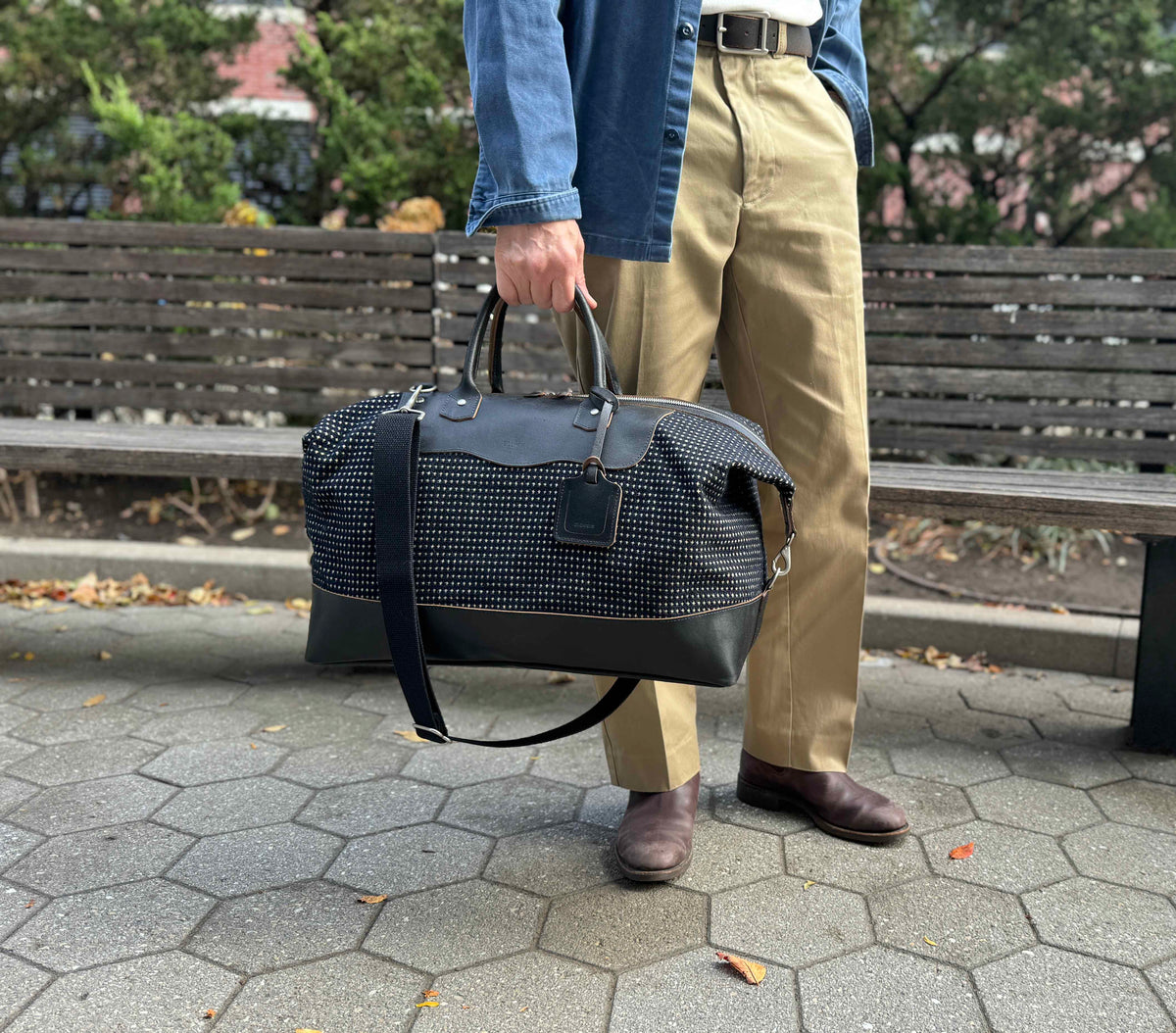 A person stands outdoors on a stone path holding a large black-and-white patterned duffel bag. They wear tan pants, brown shoes, a white belt, and a blue shirt. Wooden benches and greenery are in the background.
