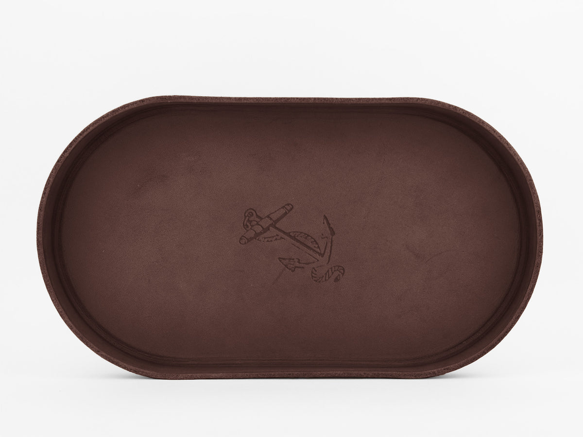 A brown, oval-shaped leather tray with a subtle engraving of an anchor in the center, set against a plain white background.
