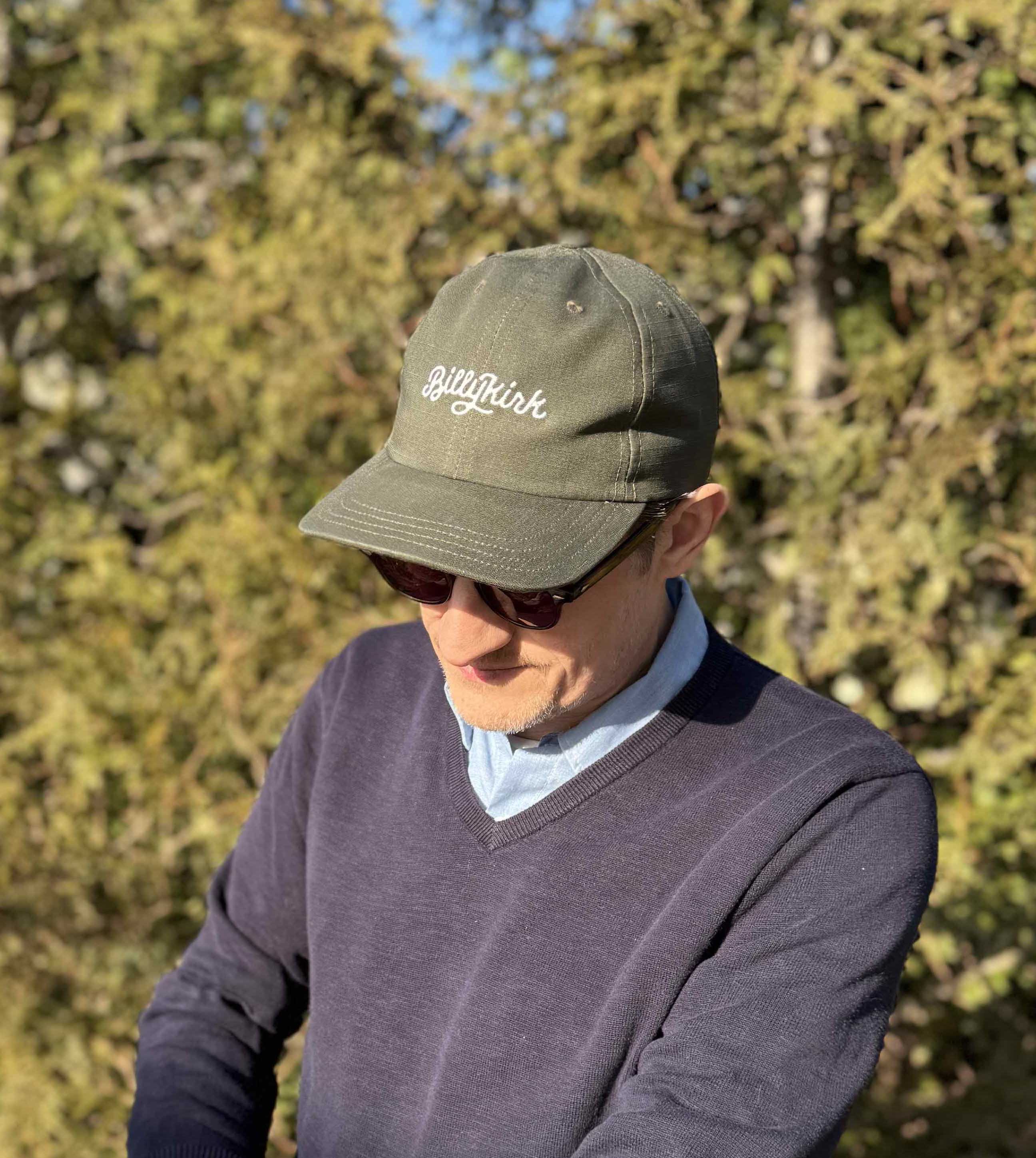 A person wearing sunglasses, an olive baseball cap, and a blue v-neck sweater stands in front of trees, looking down.