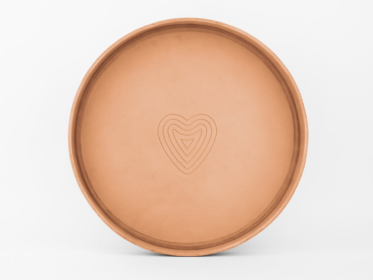 A round, shallow, tan-colored ceramic tray with a simple, embossed concentric heart design in the center, set against a plain white background.