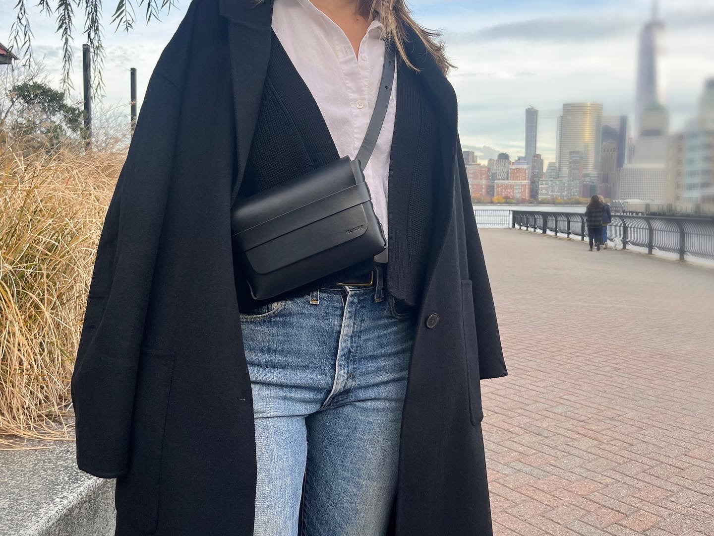 A woman wearing a long black coat, white shirt, black sweater, blue jeans, and a leather crossbody bag stands on a paved path with a city skyline and river in the background.