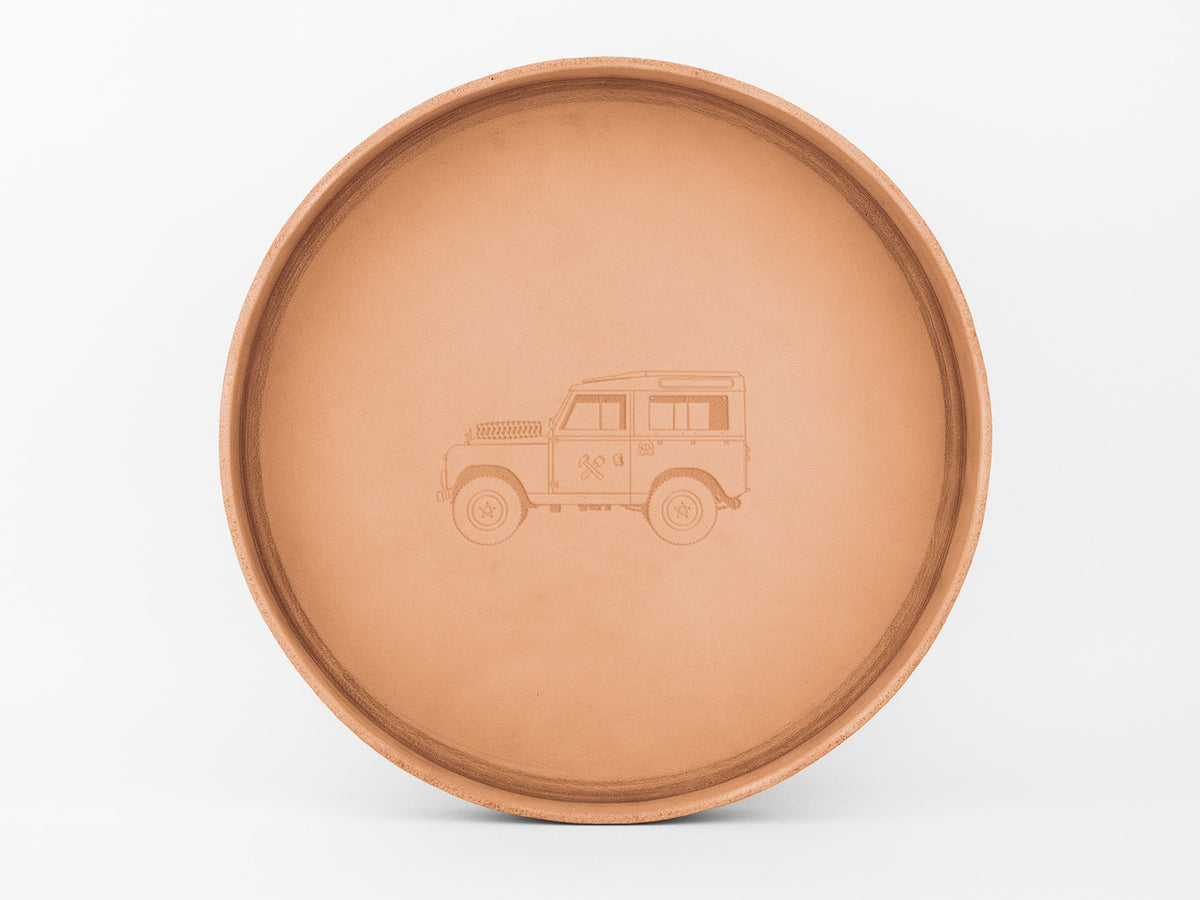 A round, light brown ceramic plate with an engraved illustration of a classic off-road vehicle in the center, set against a plain white background.