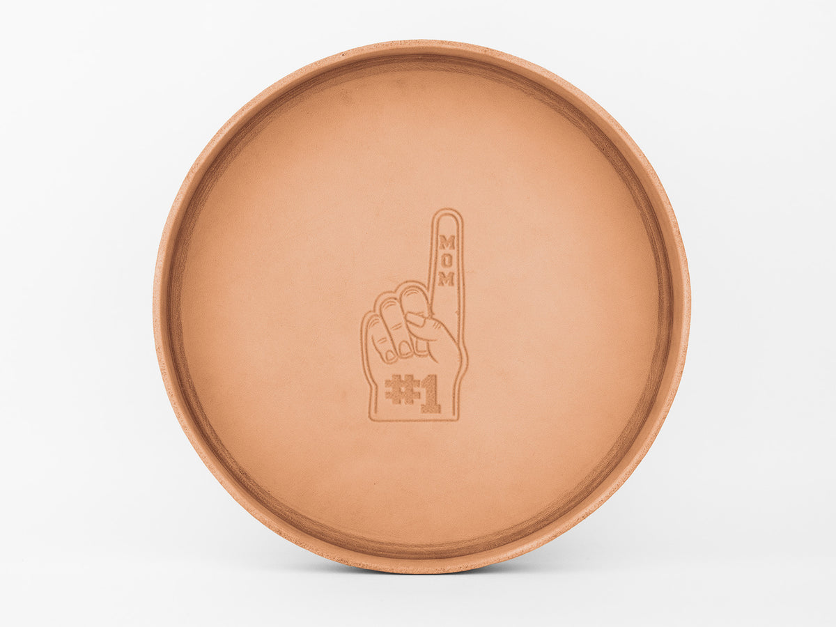 A round, tan tray with an engraved image of a foam finger. The finger points up and has MOM written vertically on the finger and #1 at the base, symbolizing Number 1 Mom. The tray is on a white background.