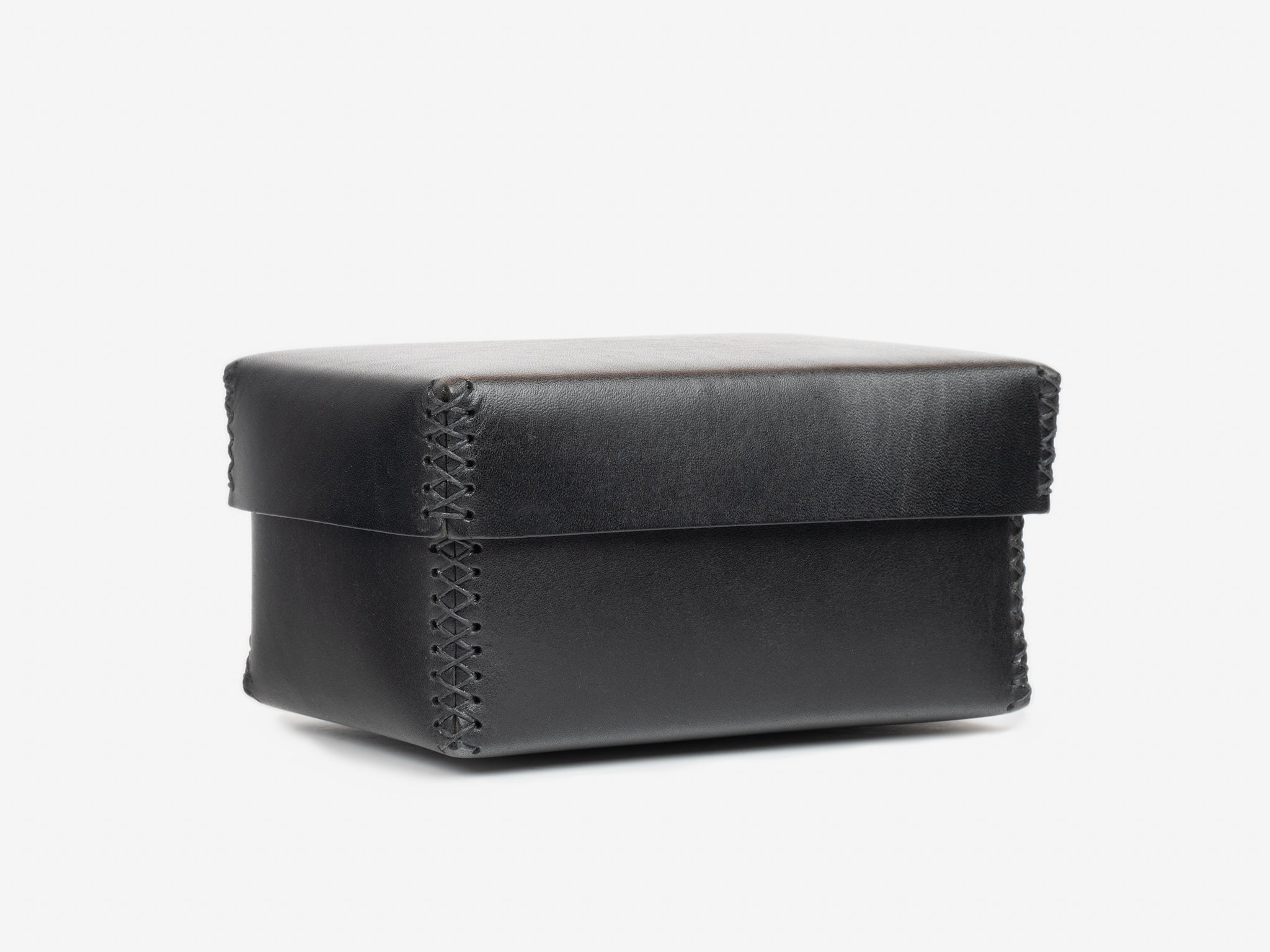 A black, rectangular leather box with visible stitching along the edges, set against a plain white background. The box has a fitted lid and a smooth, minimalistic design.