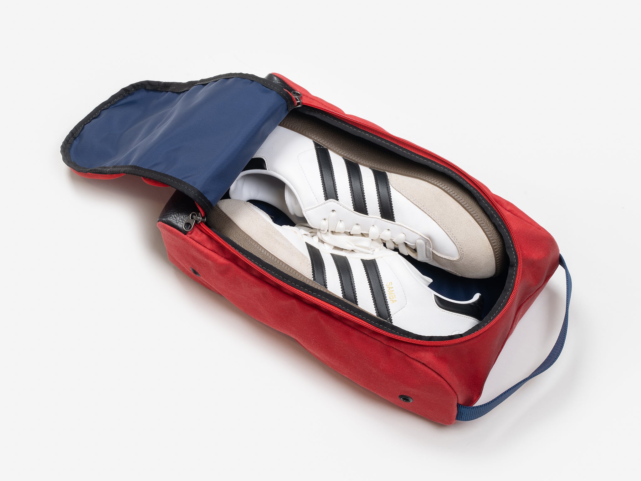 A pair of white sneakers with black stripes placed inside a red shoe bag with a navy blue interior and the top flap partially unzipped.