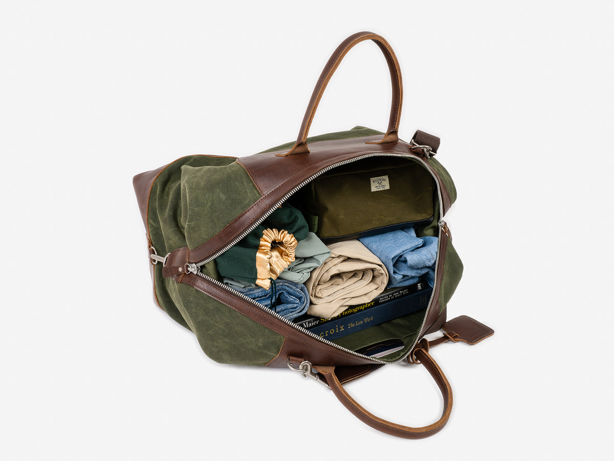 A Green and Brown patterned duffel bag with brown handles sits open, revealing neatly folded clothes, a toiletry bag, and a blue book inside, against a plain white background.
