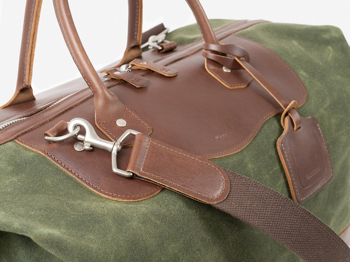 A close-up of a Green and Brown duffel bag with leather handles, silver zipper, and a detachable shoulder strap; the bag also features metal hardware and a leather luggage tag.