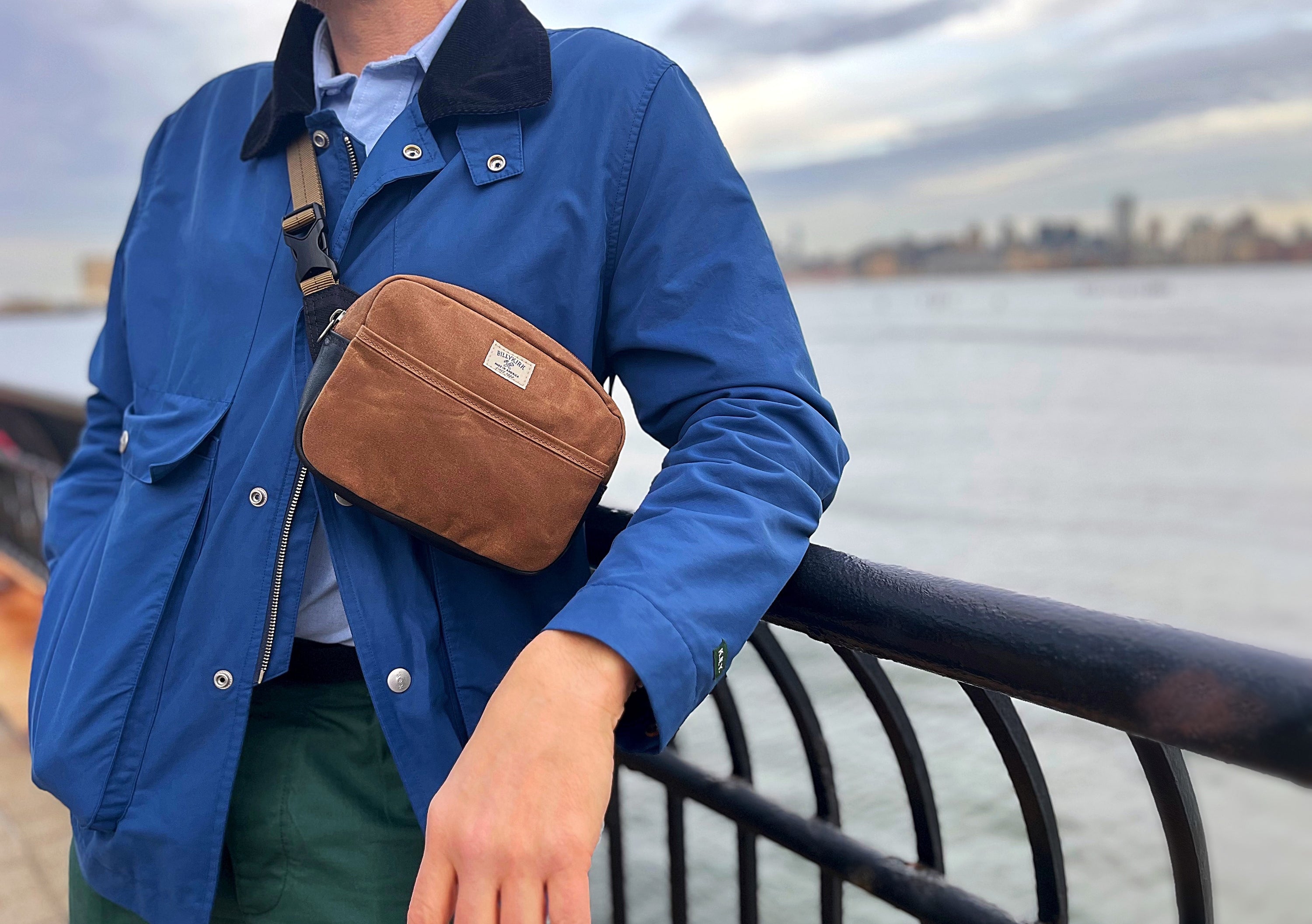 A person wearing a blue jacket and green pants stands by a waterfront railing, with a brush brown waxed canvas crossbody bag across their chest. The background shows water and a distant city skyline under a cloudy sky.