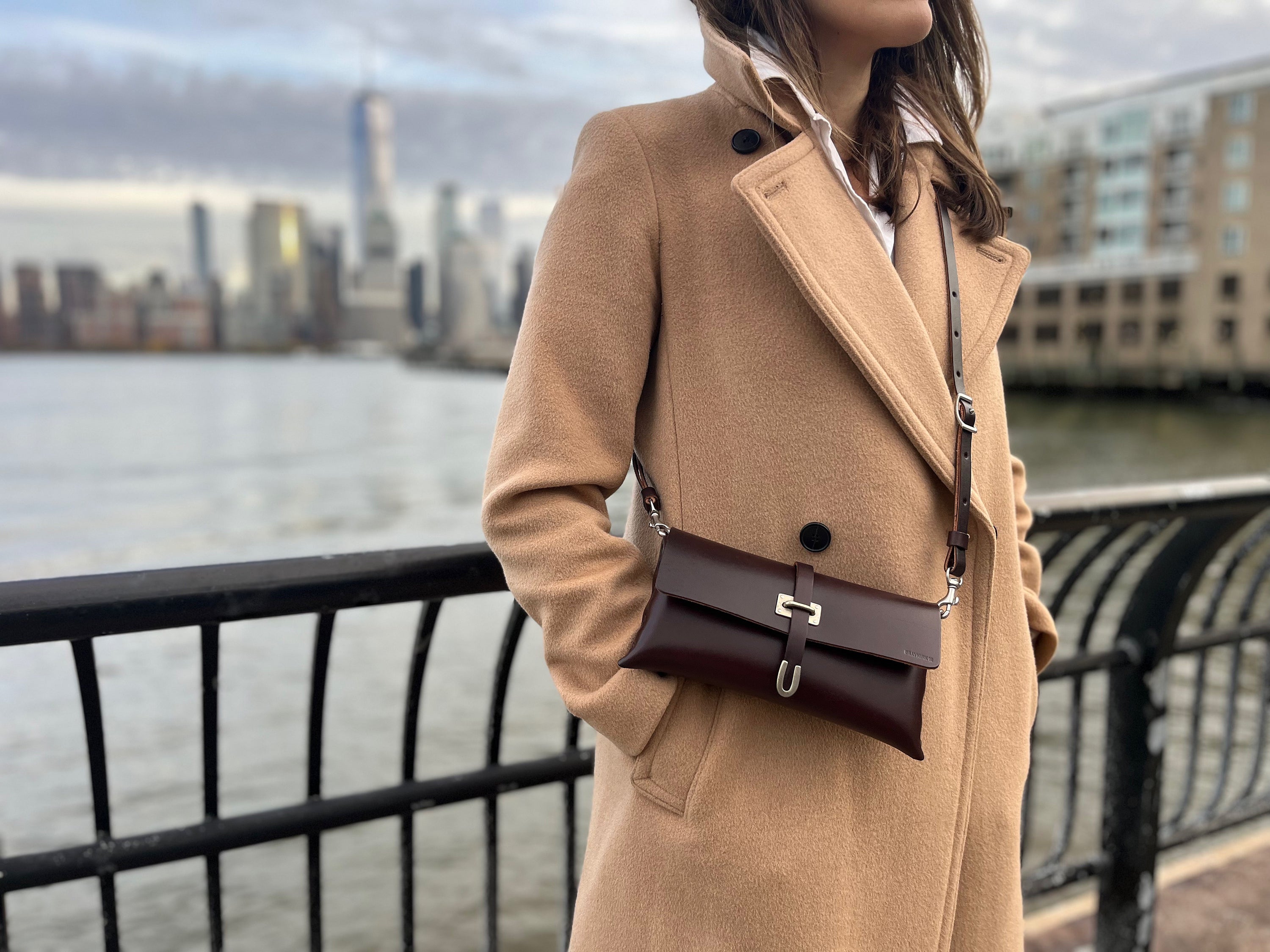 A woman in a tan coat with a brown crossbody purse stands by a waterfront railing, with a city skyline and tall buildings blurred in the background.