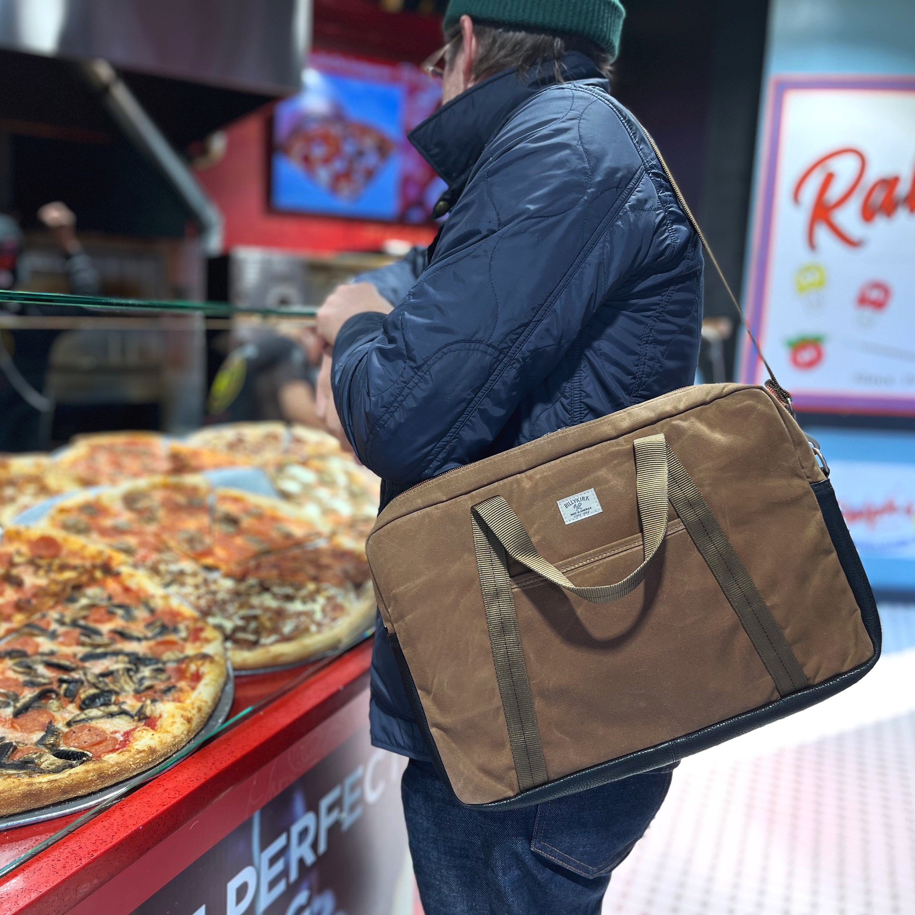 A person in a navy jacket and green beanie carries a waxed canvas brown briefcase on his shoulder while standing at a counter with pizzas on display.