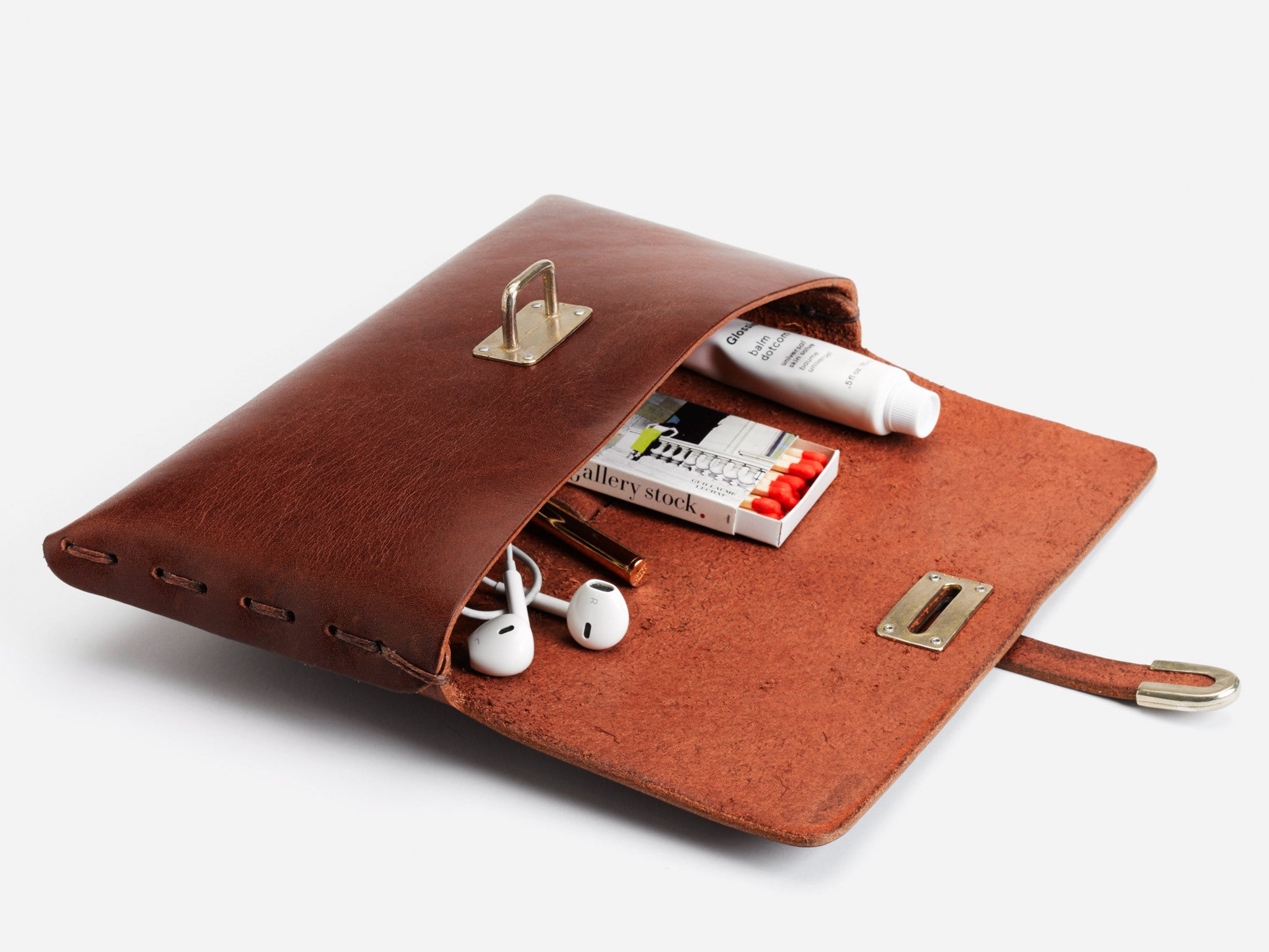 A brown leather pouch lies open, displaying its contents: white earphones, pens, a small notepad, a white tube, and a pack of colored pencils against a white background.