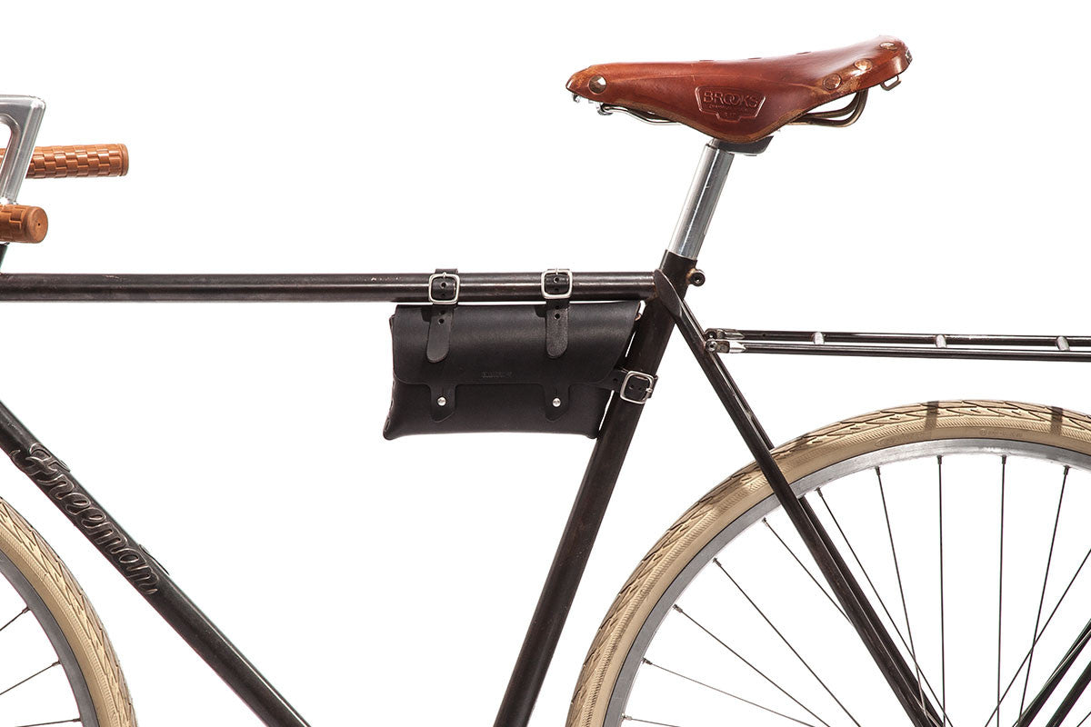 Side view of a black bicycle with tan tires, a brown leather saddle, and a small black leather tool bag attached to the frame below the seat. The background is white and uncluttered.