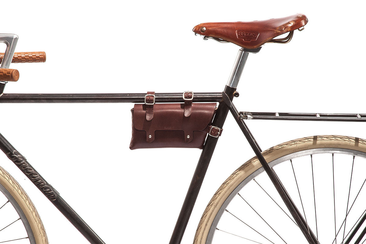 A close-up side view of a vintage-style bicycle with a brown leather seat, beige tires, and a brown leather bag attached to the frame beneath the seat. The background is white.