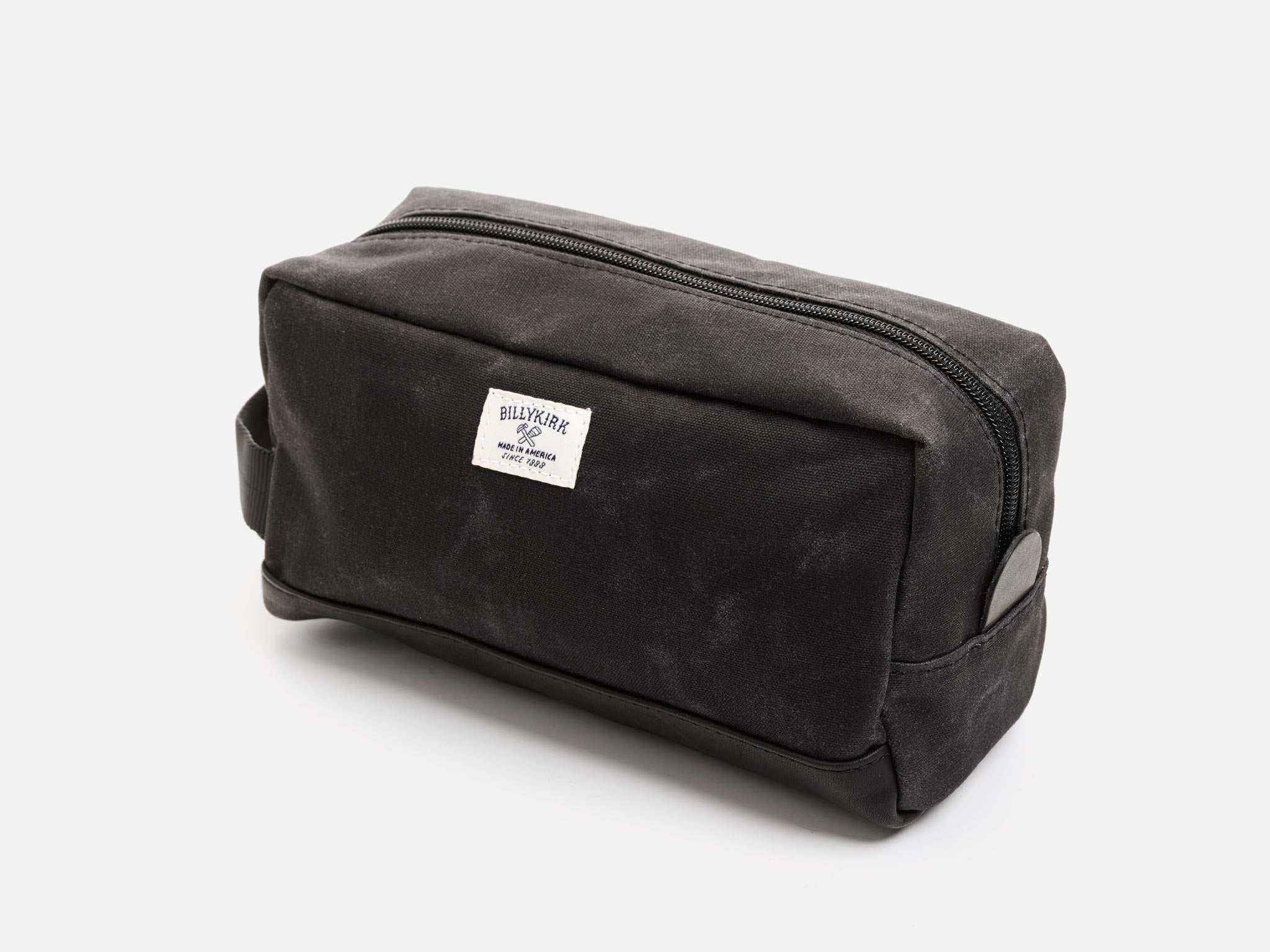 A rectangular black waxed canvas toiletry bag with a zipper, a black nylon webbing handle, a black leather base, a leather tab to help close the zipper, and an off-white Billykirk brand label on the front.