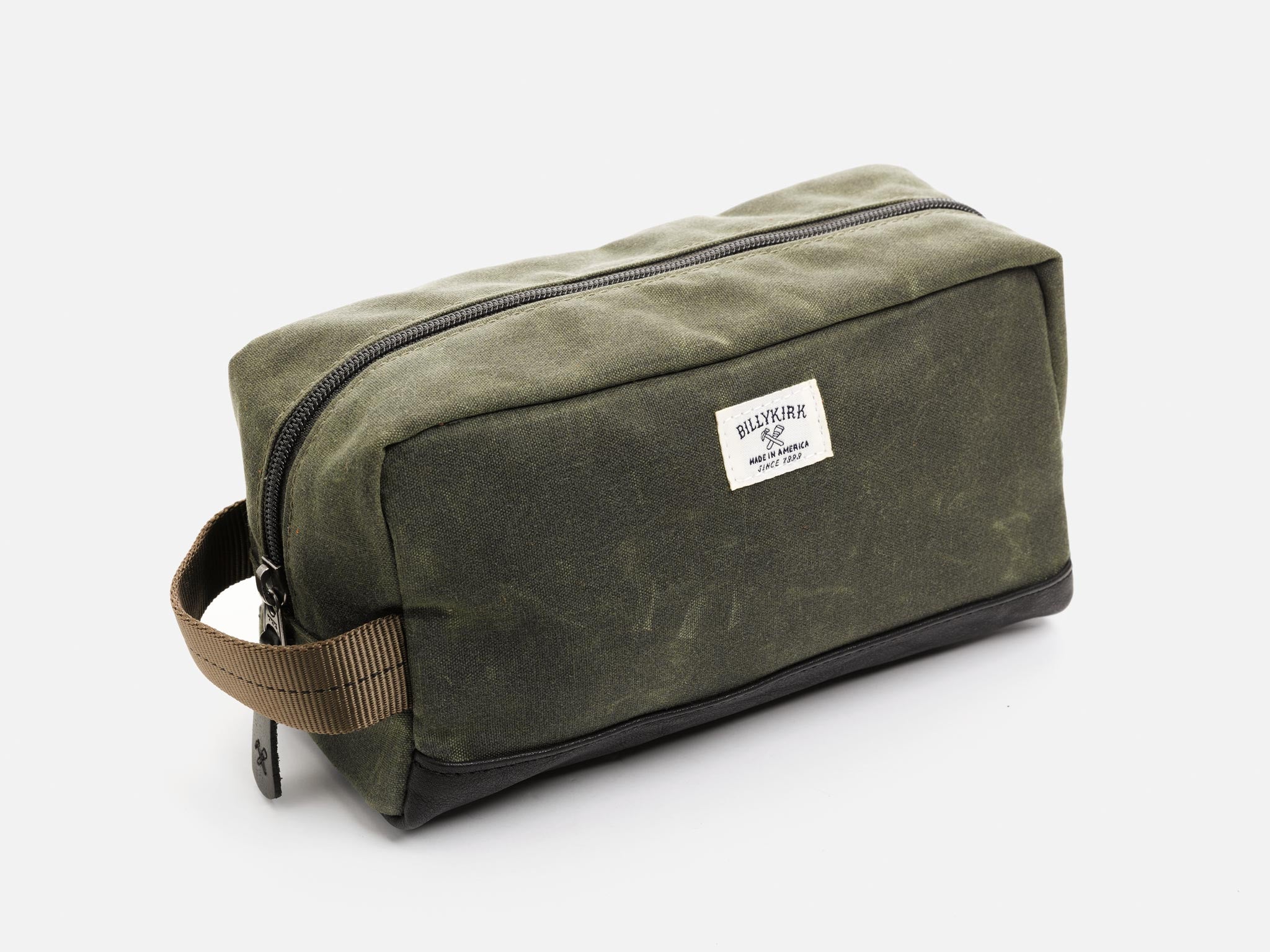 A rectangular olive waxed canvas toiletry bag with a zipper, a black nylon webbing handle, a black leather base, and an off-white Billykirk brand label on the front.