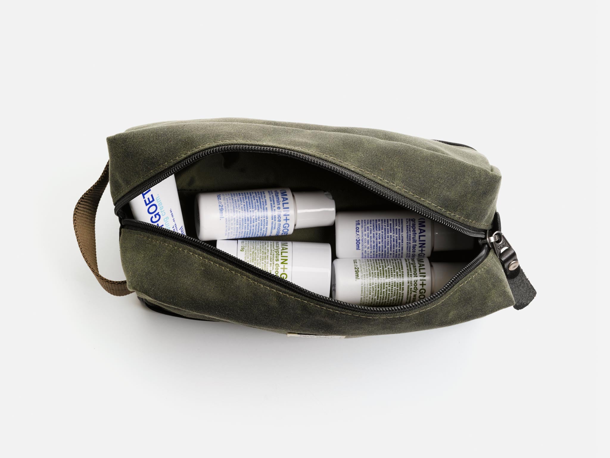 An olive waxed canvas toiletry bag unzipped to reveal several Malin+Goetz branded bottles and containers inside, arranged neatly against a white background.
