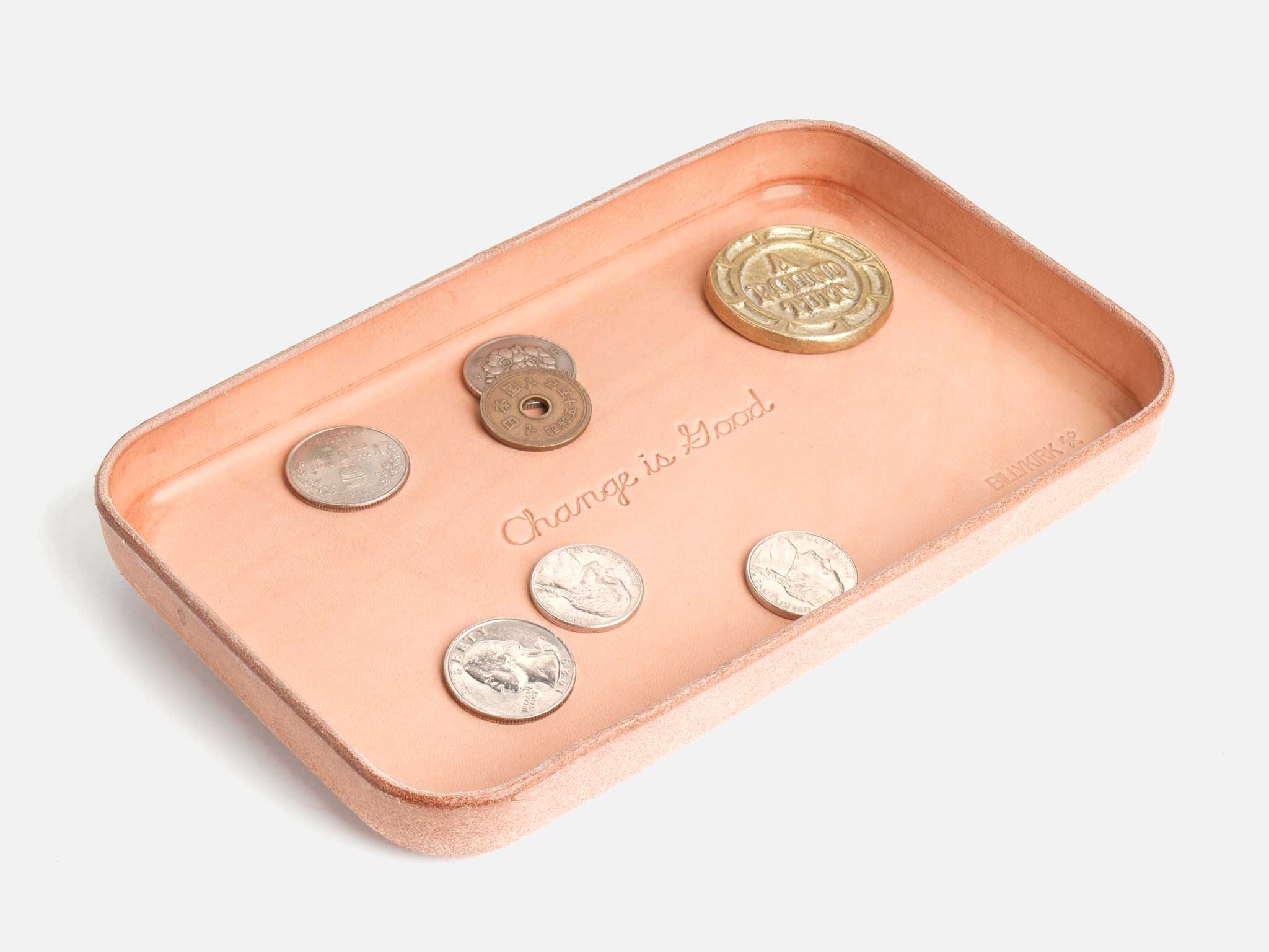 A tan leather tray holds assorted coins. The phrase Change is Good is embossed in the center of the tray. The background is plain white.