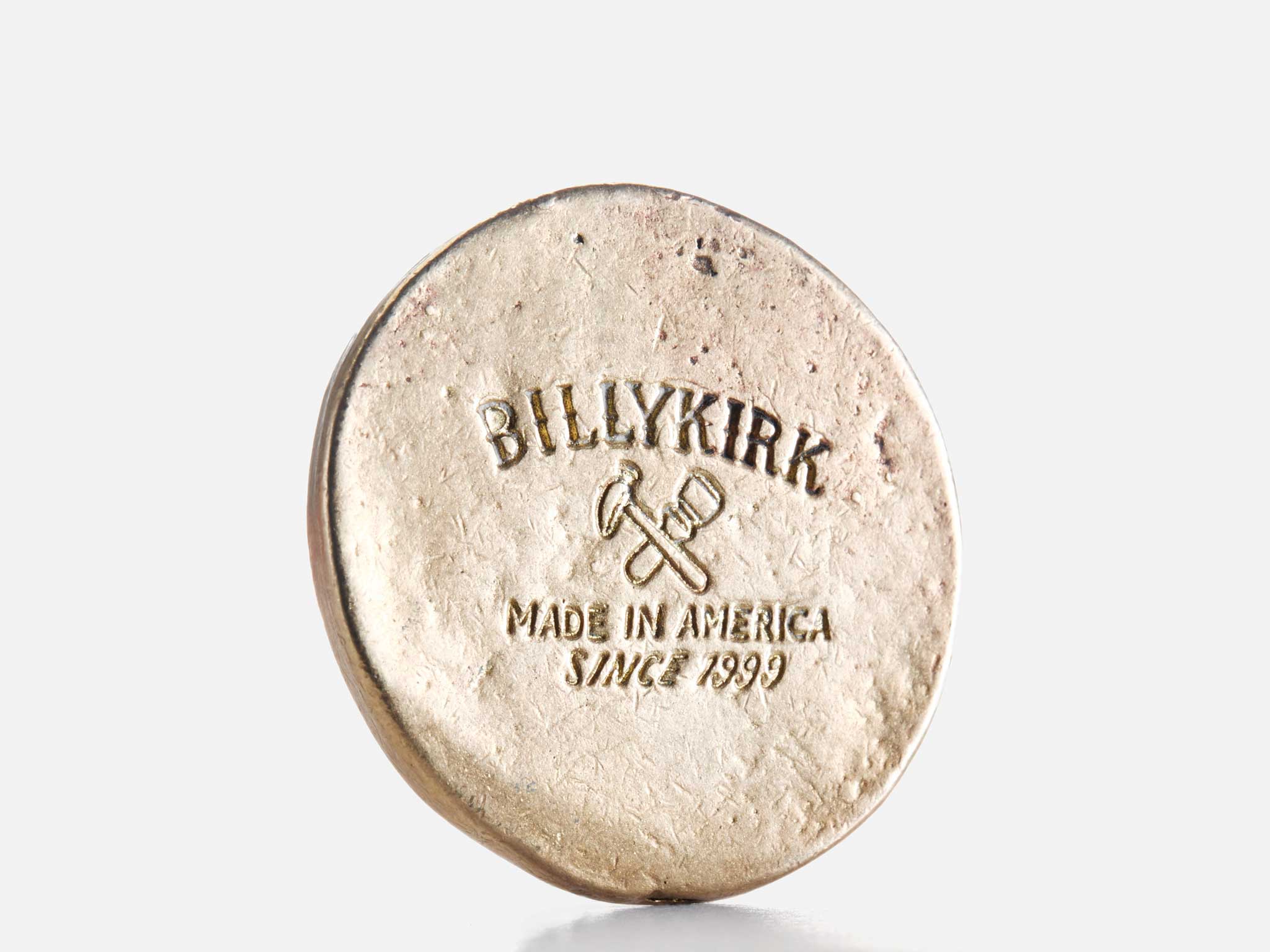 A round, metal object is stamped with the text BILLYKIRK MADE IN AMERICA SINCE 1999 and an image of two crossed tools, set against a plain white background.
