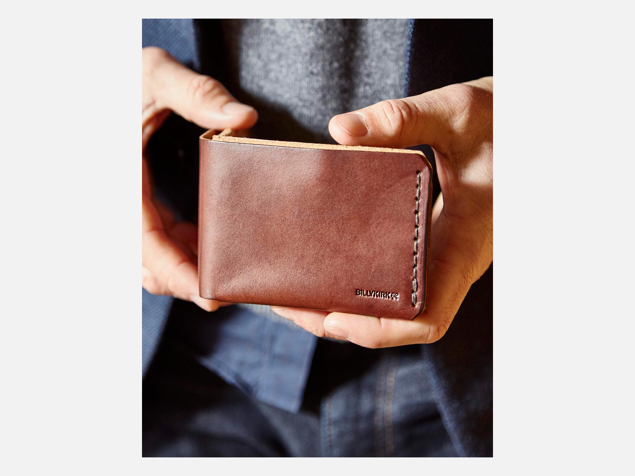 A person holds a brown leather wallet with visible stitching and the word BELLROY embossed in the lower corner. The person is wearing a dark jacket and gray shirt.