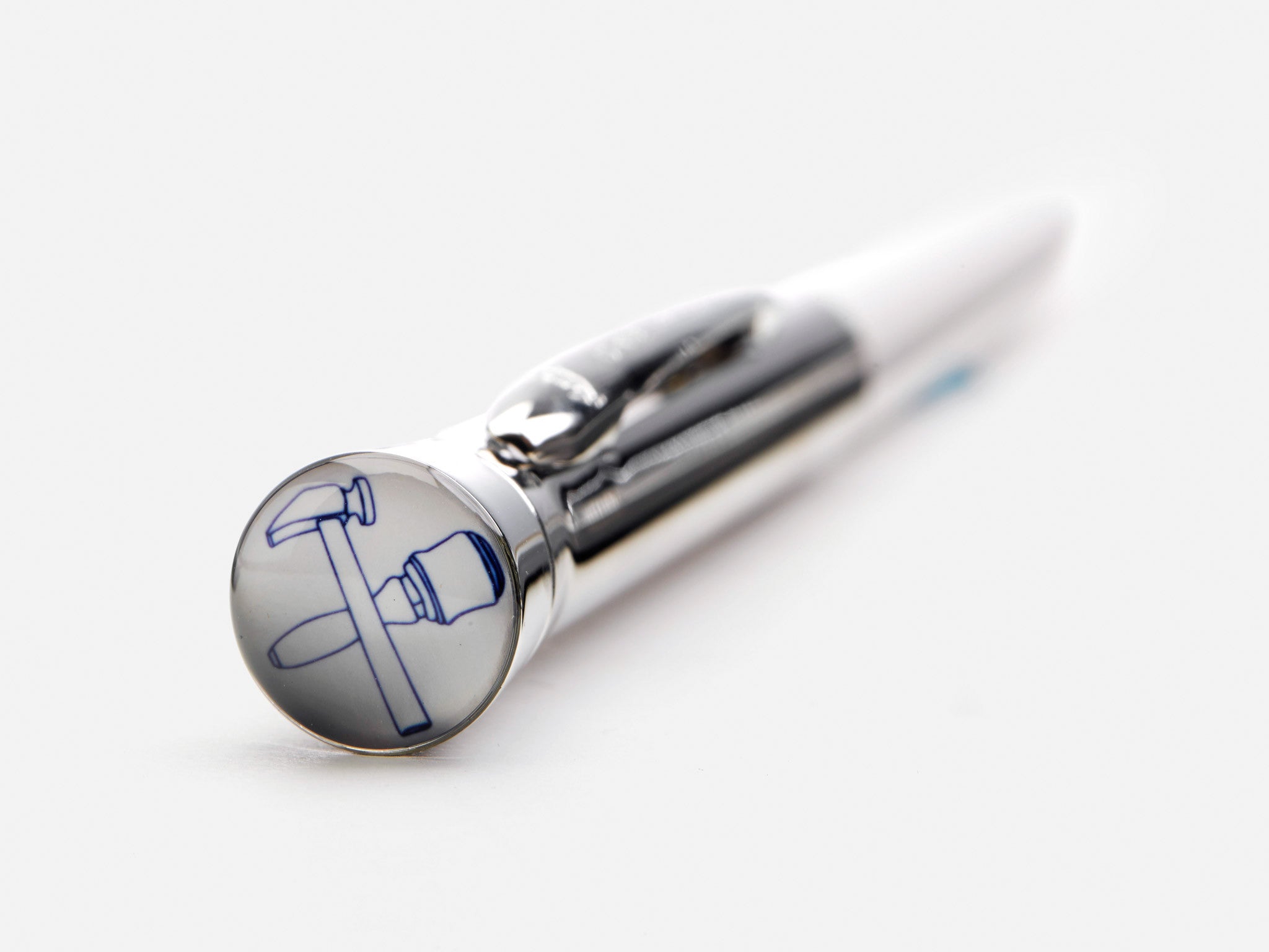 A close-up of a metallic pen resting on a white surface, with a round emblem featuring a blue hammer and pickaxe crossed on the end of the pen.