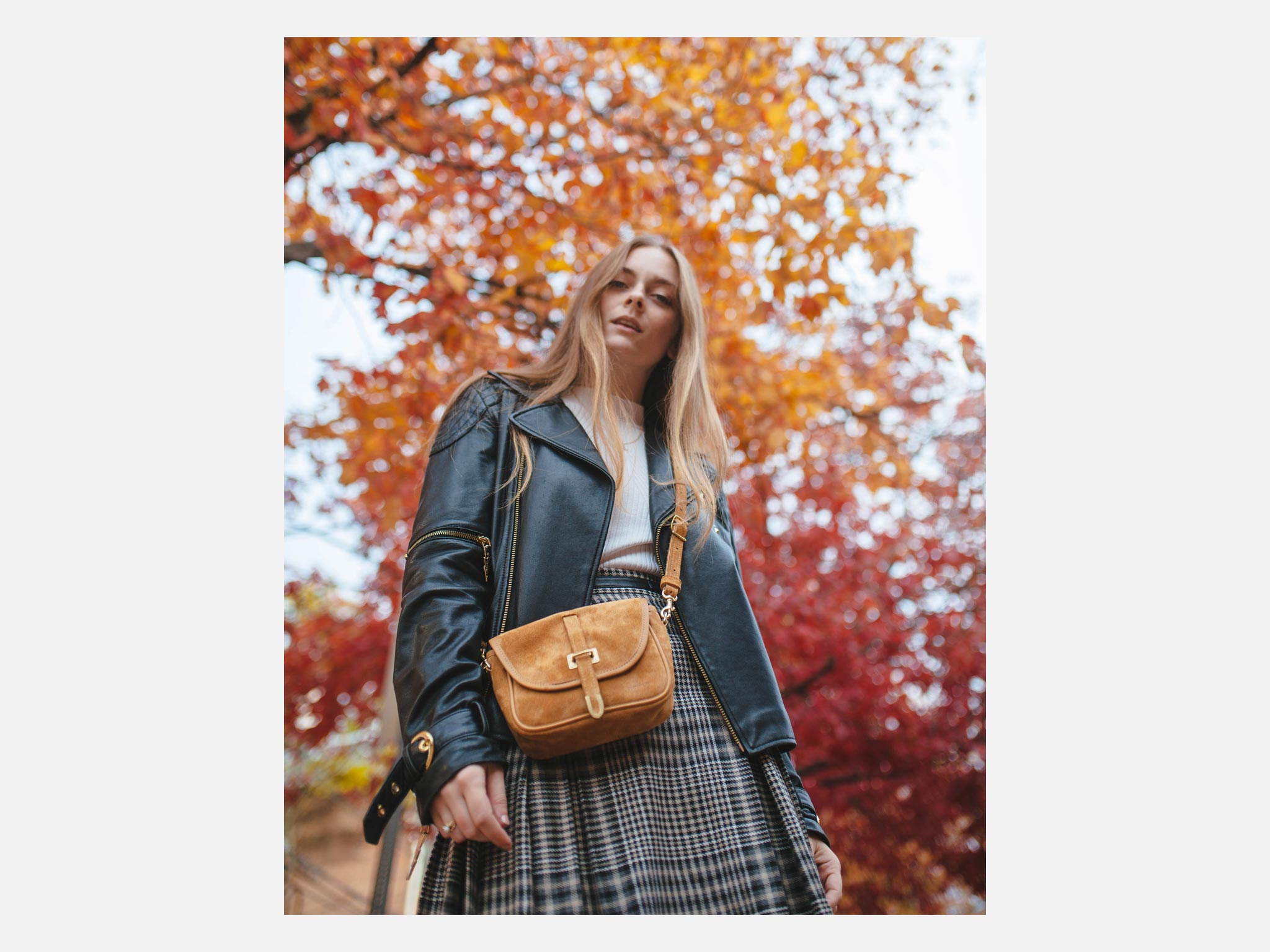A woman in a black leather jacket and plaid skirt stands outdoors with autumn trees in the background, holding a small tan crossbody bag. The leaves are red and orange, creating a vibrant fall atmosphere.