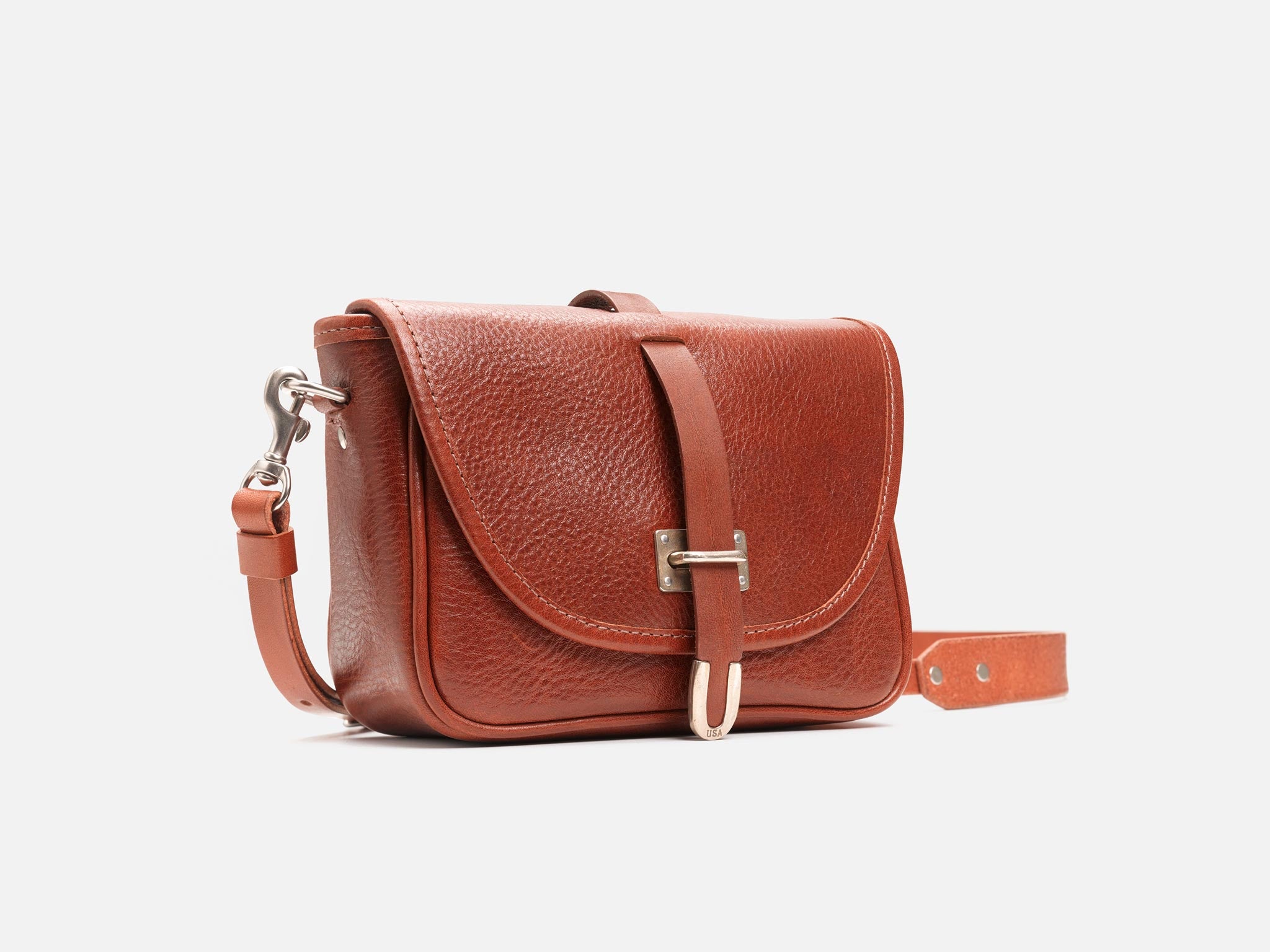 A small, brown leather crossbody bag with a textured finish, flap closure, silver buckle, and adjustable strap, displayed against a plain white background.