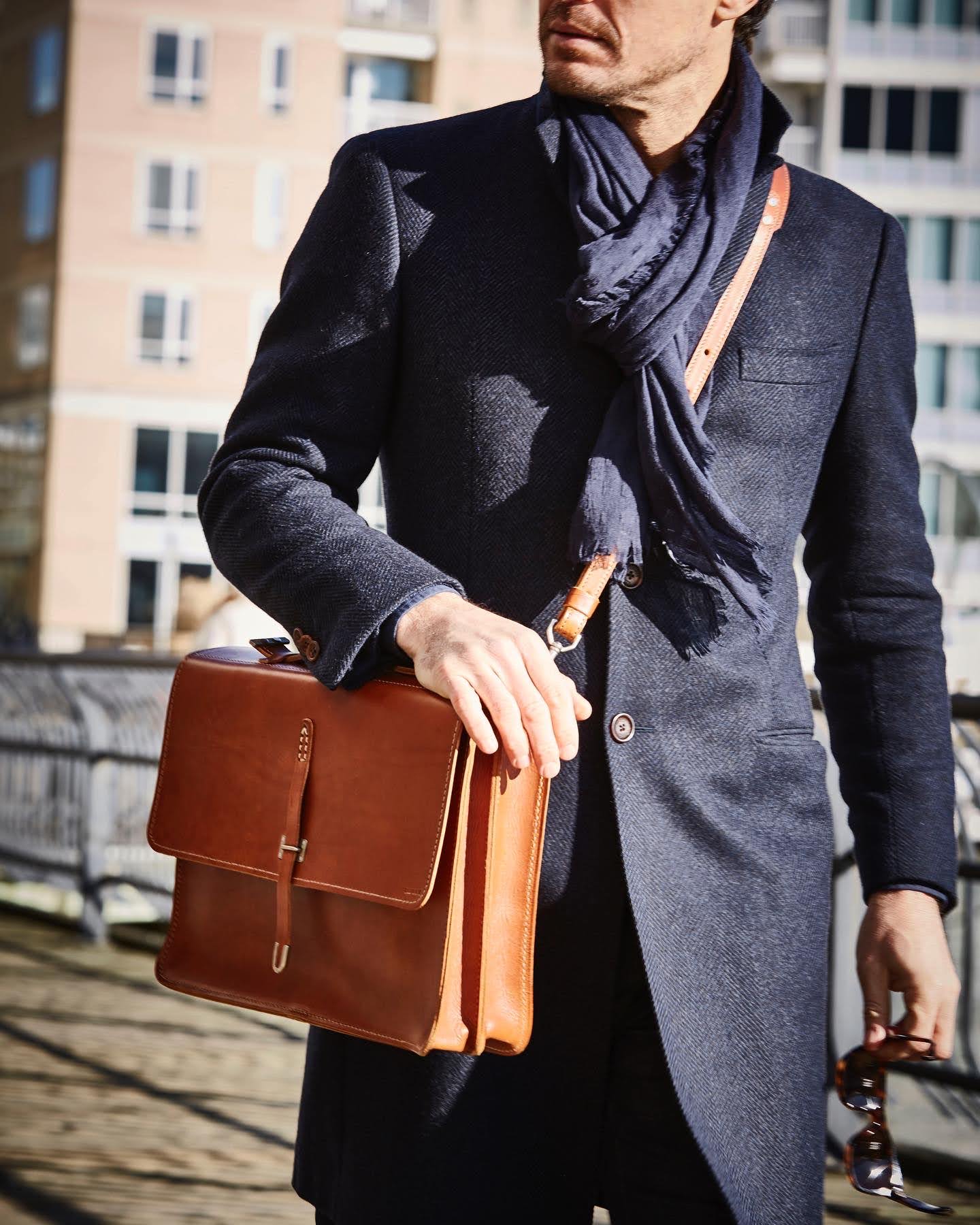A man in a dark coat and scarf carries a leather briefcase over his shoulder and holds sunglasses. Urban buildings are blurred in the background.