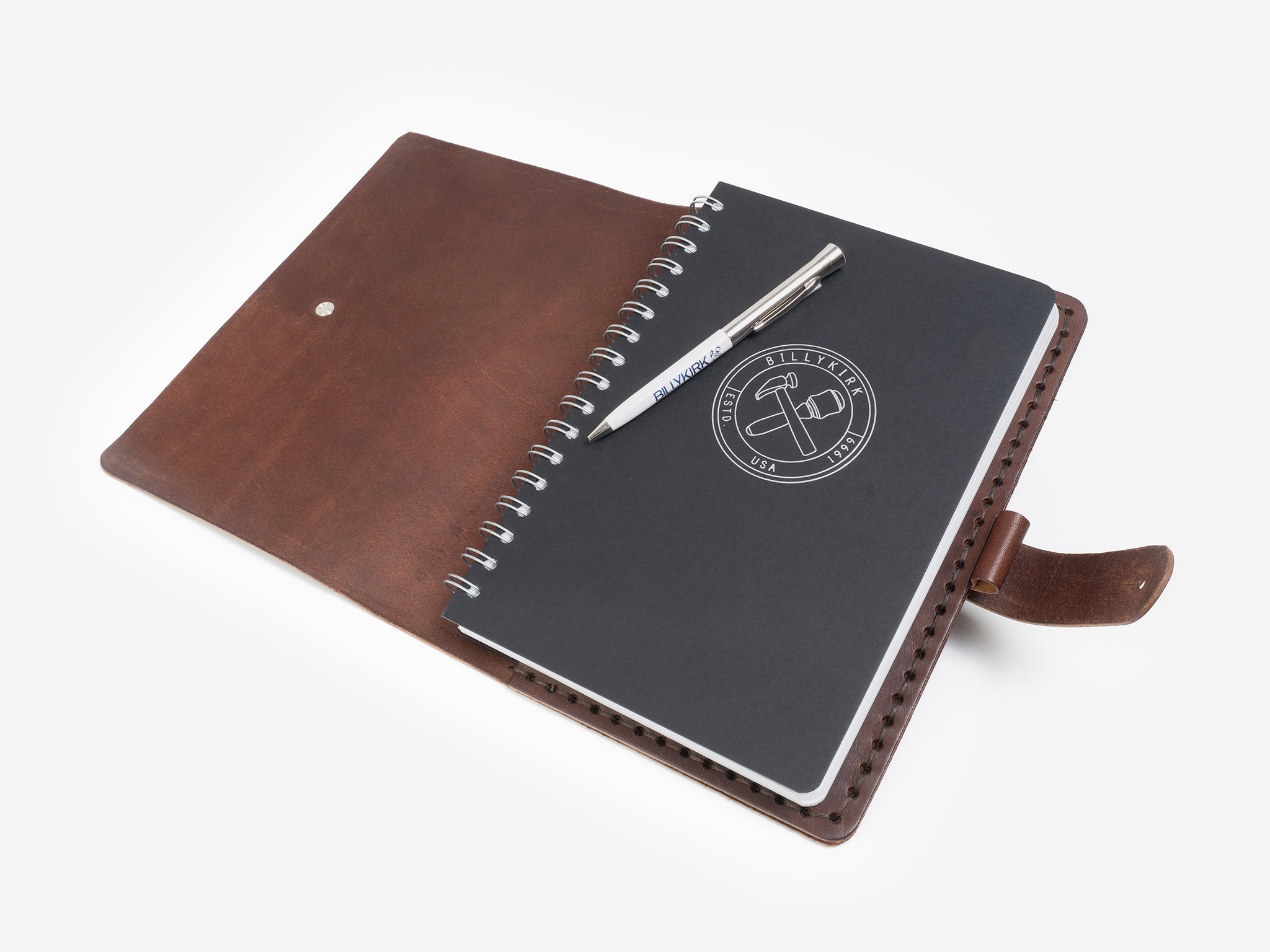 A brown leather binder lays open on a white surface, holding a spiral-bound notebook with a dark cover and a logo. A silver and blue pen rests diagonally across the notebook.