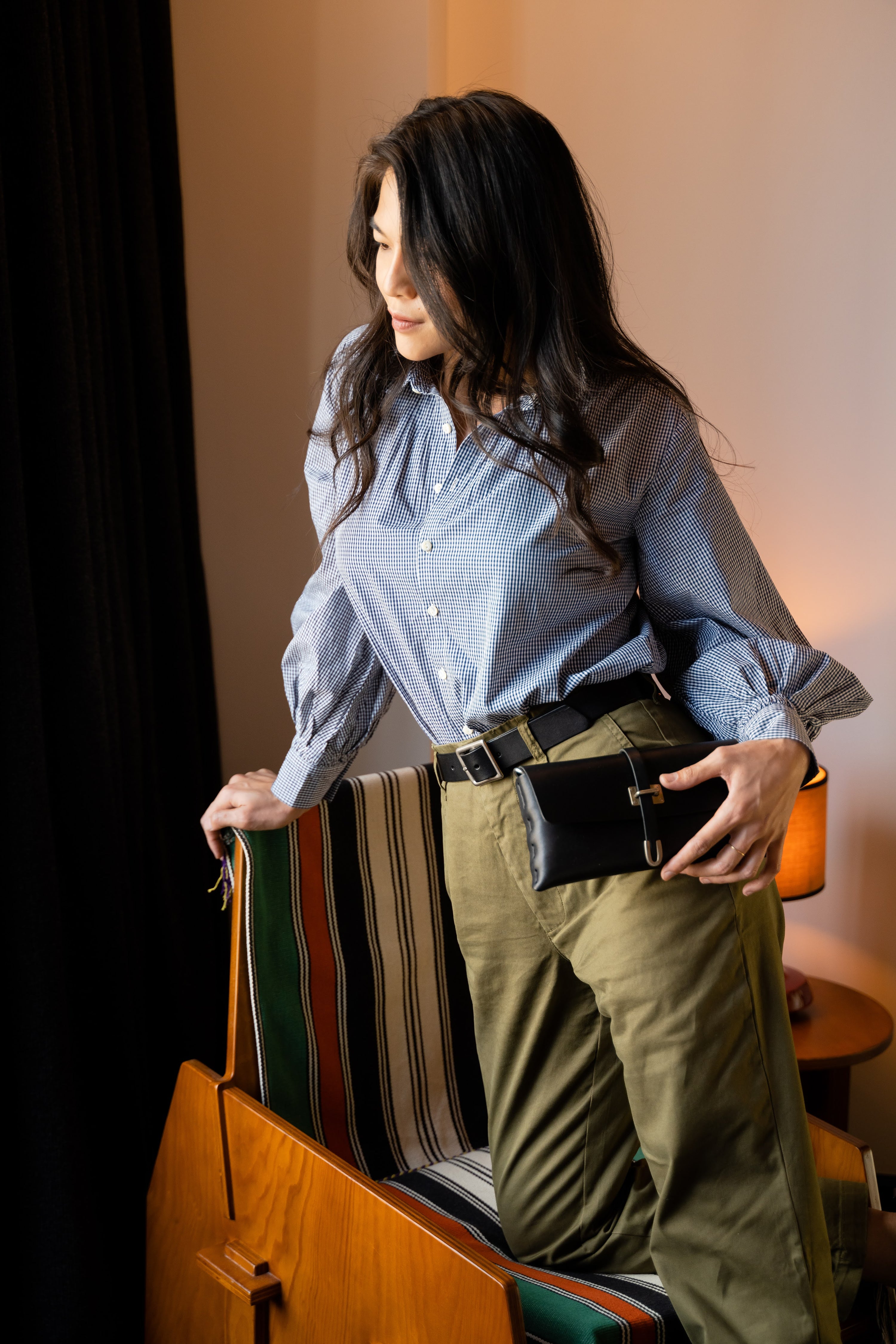 A woman with long dark hair stands next to a striped chair, wearing a blue checkered blouse and olive green pants with a black belt purse, looking down with a relaxed expression in a softly lit room