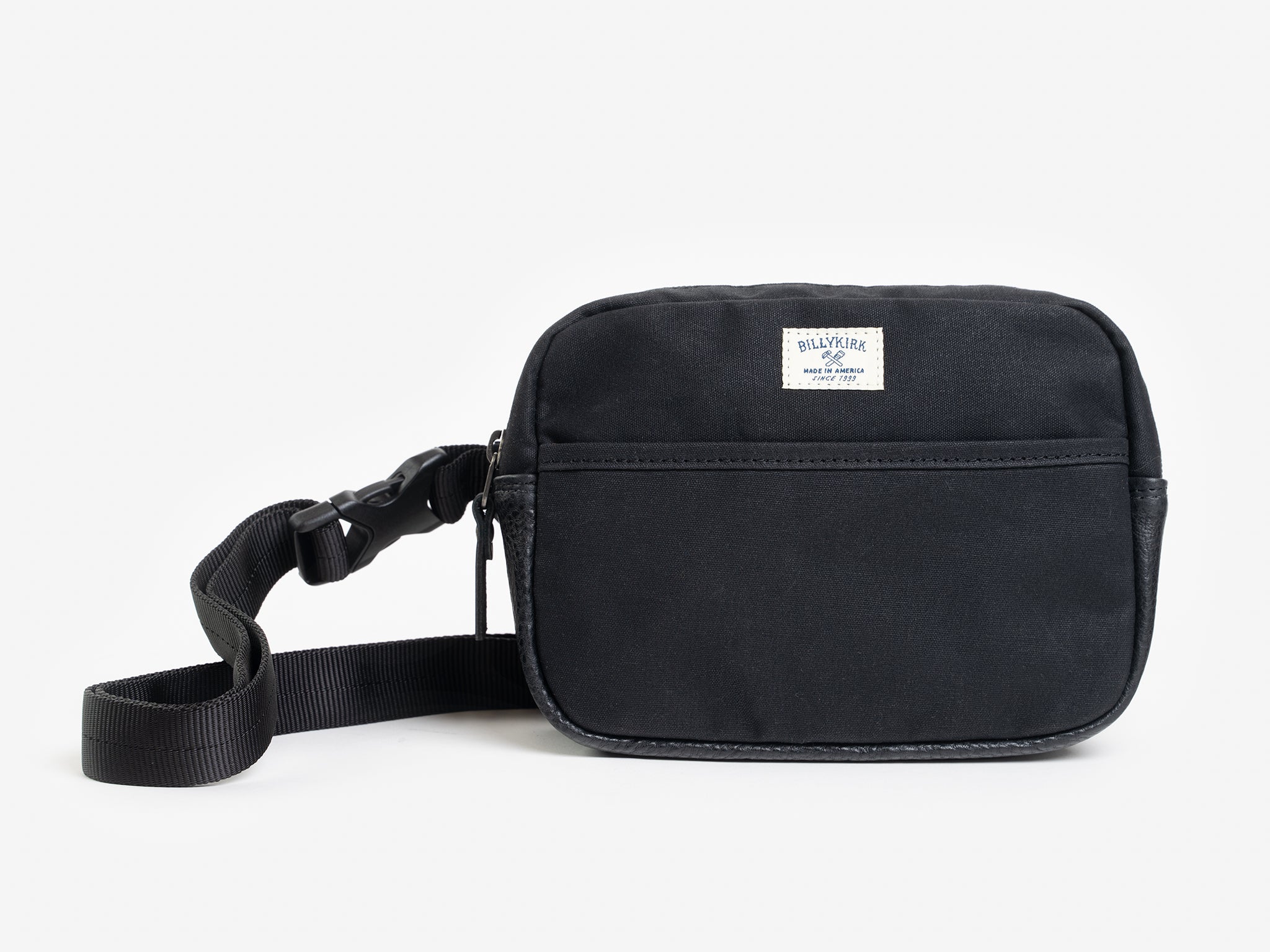 A small black waxed canvas crossbody bag with a front pocket, adjustable strap, and a cream colored rectangular Billykirk brand patch stitched on the front.