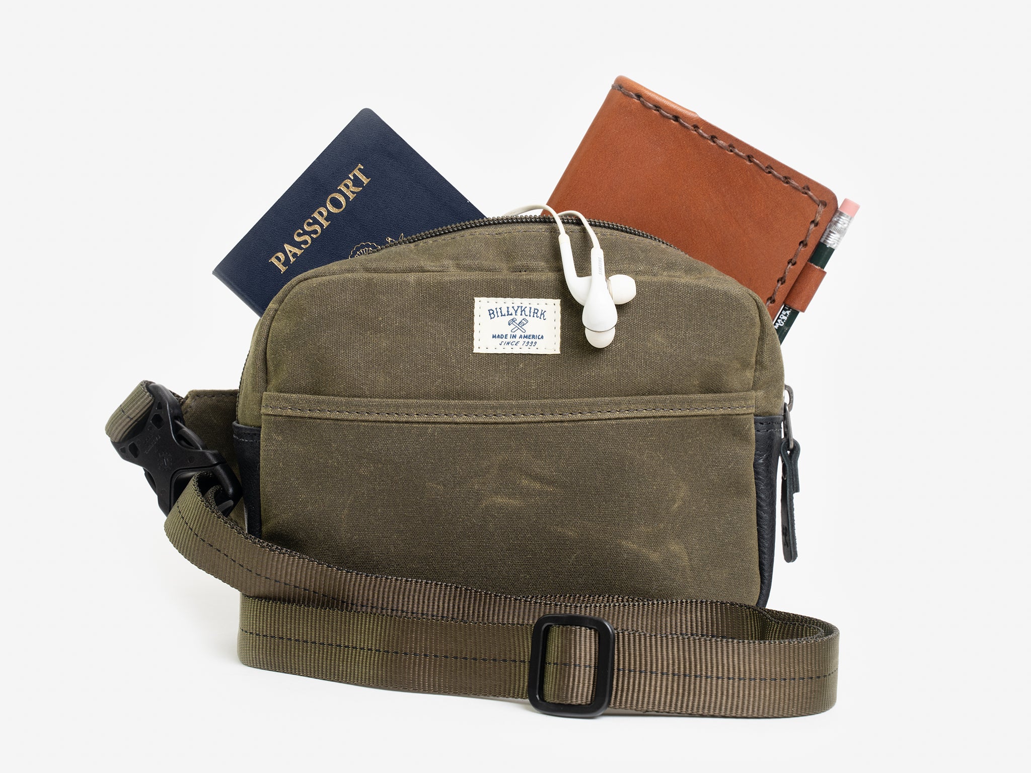A small olive waxed canvas crossbody bag with a wallet, passport, and white earphones sticking out of the zippered top. The bag has a front pocket, and a cream colored rectangular Billykirk brand patch stitched on the front. 