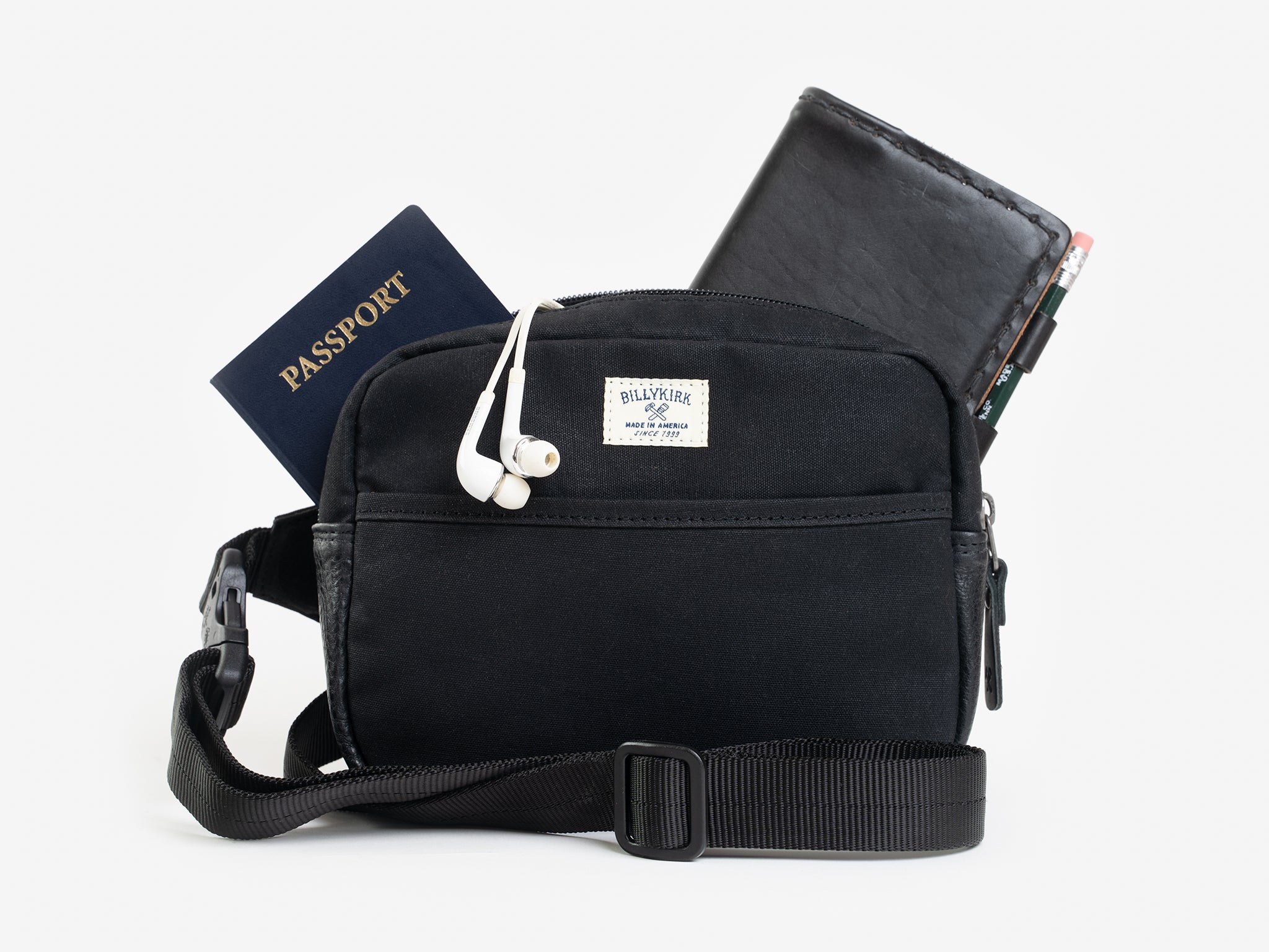 A small black waxed canvas crossbody bag with a wallet, passport, and white earphones sticking out of the zippered top. The bag has a front pocket, and a cream colored rectangular Billykirk brand patch stitched on the front. 
