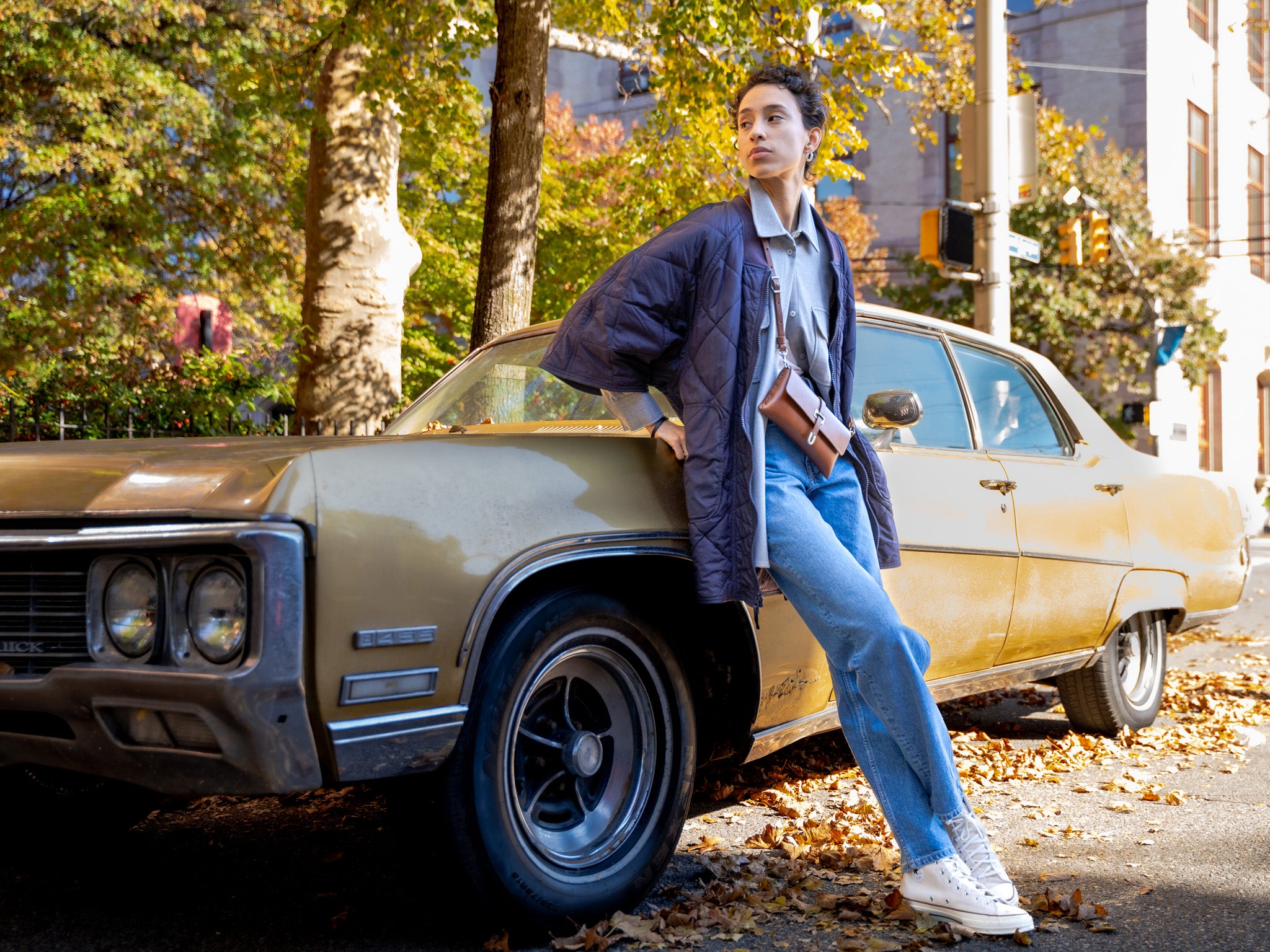A person in jeans, a blue shirt, and a jacket leans against an old gold-colored car parked on a leaf-strewn street, with autumn trees and buildings in the background.