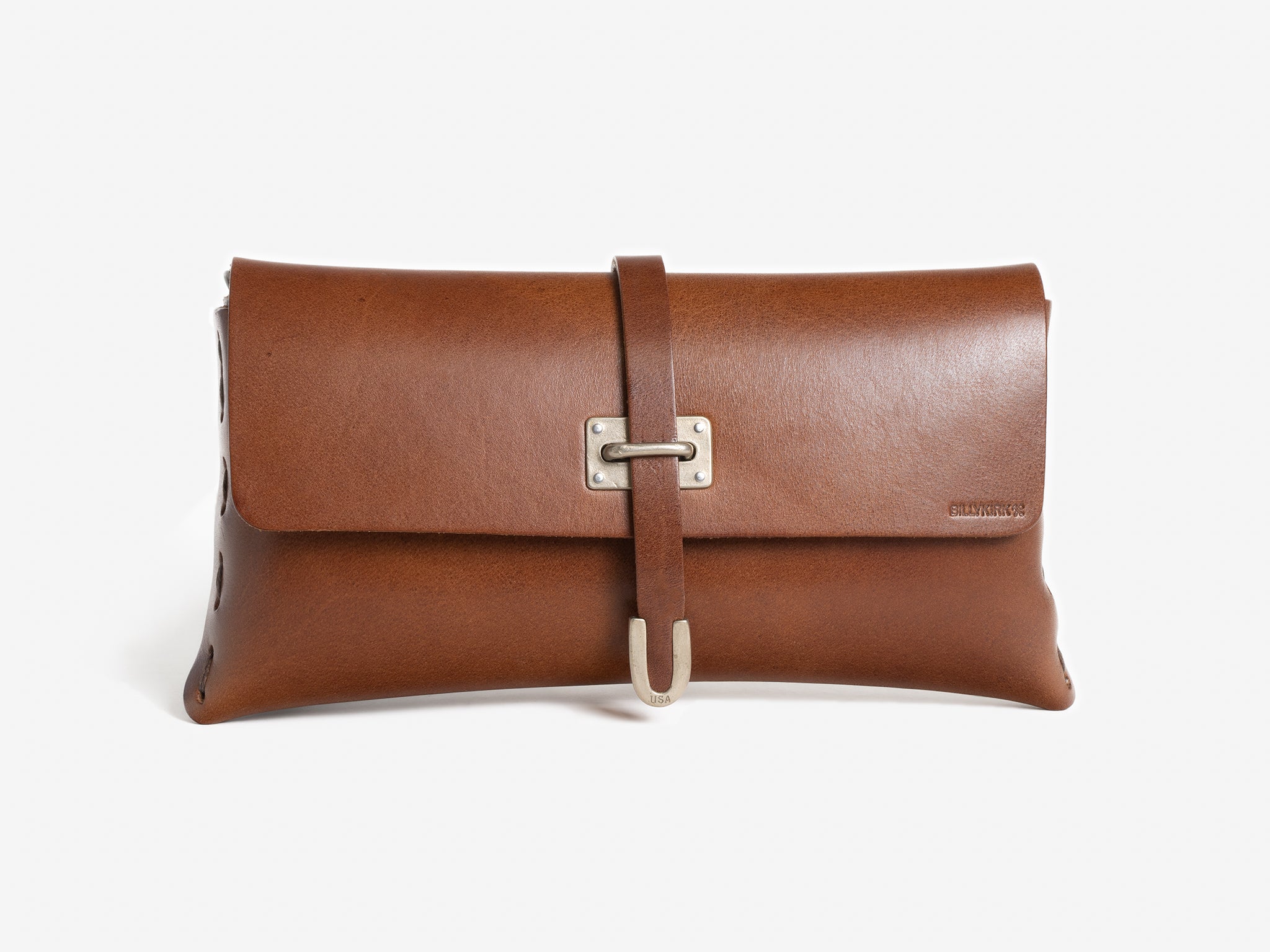A brown leather clutch bag with a front flap, secured by a strap and metal buckle. The bag has minimal stitching on the sides and a small embossed logo on the front right corner.