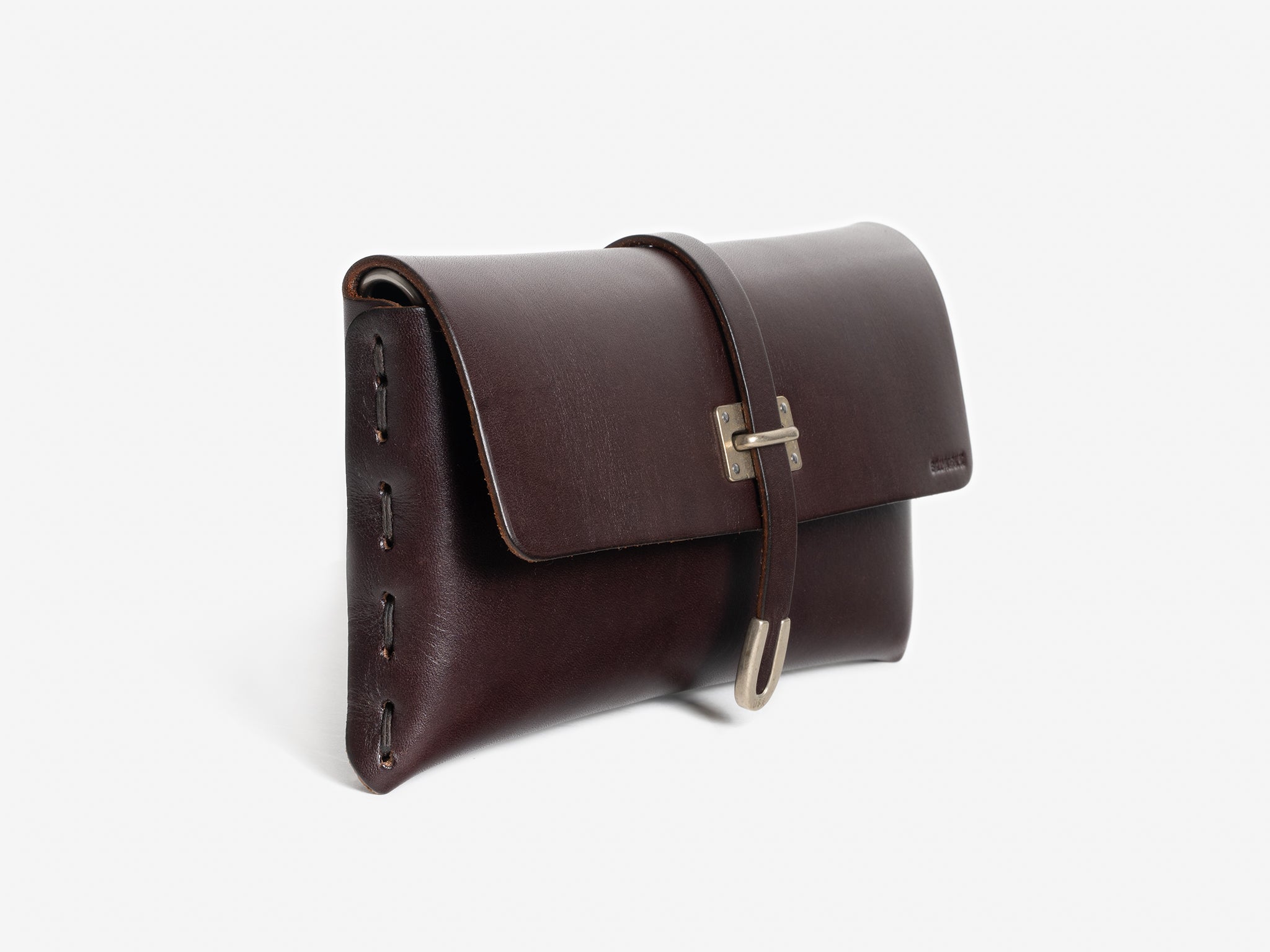 A dark brown leather clutch with a flap closure and a metal buckle, featuring side stitching and a minimalist design, set against a plain white background.