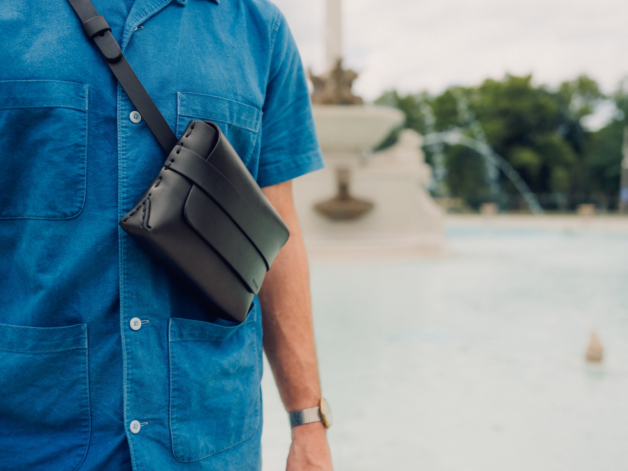A person wearing a blue button-up shirt and a black leather crossbody bag stands near a large outdoor fountain with water spraying in the background. Only their torso and one arm are visible.