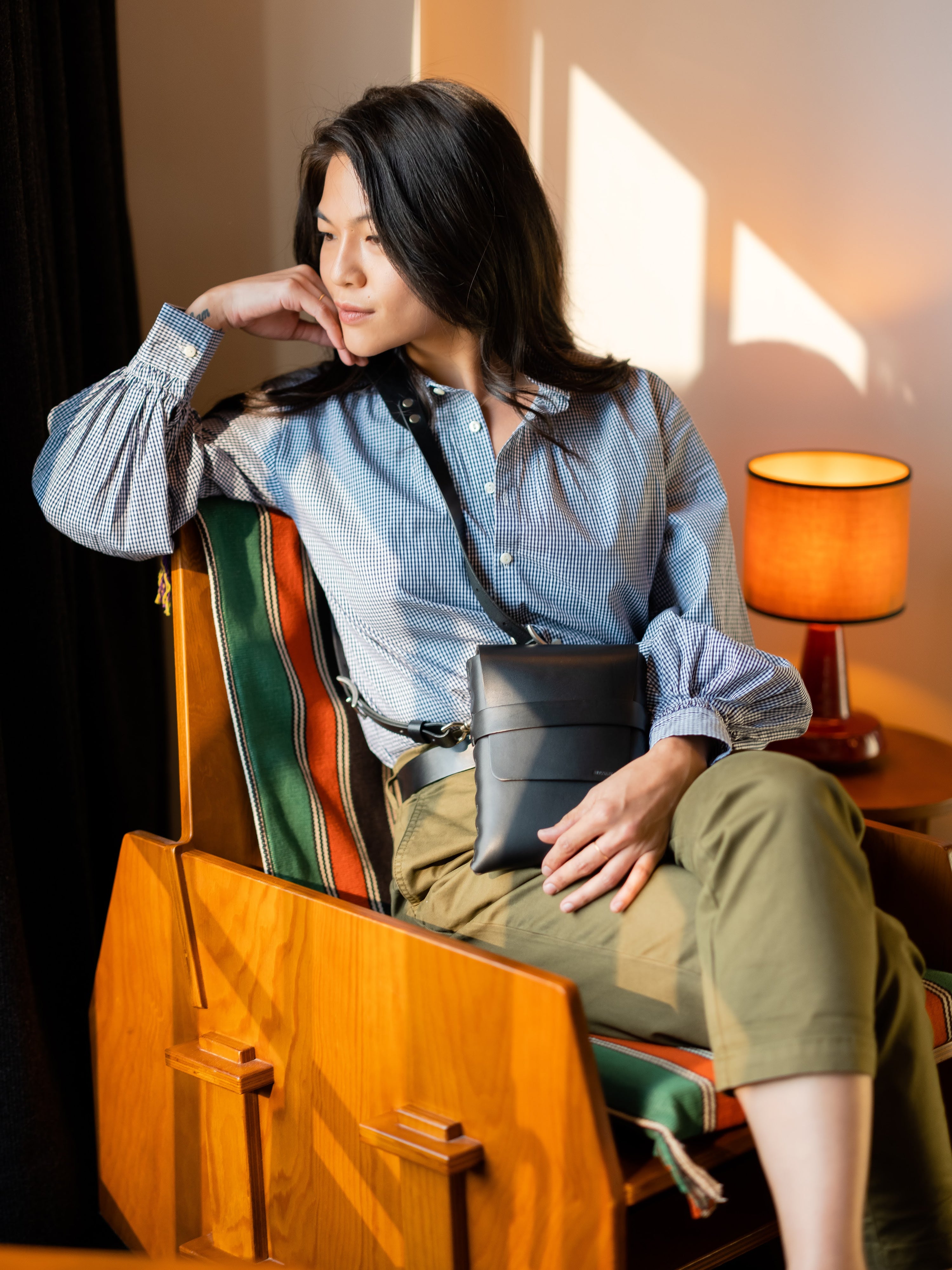 A woman with long dark hair sits on a wooden chair by a window, wearing a striped shirt and olive pants, holding a black purse, and gazing thoughtfully to the side. Warm sunlight and a lit lamp brighten the scene.