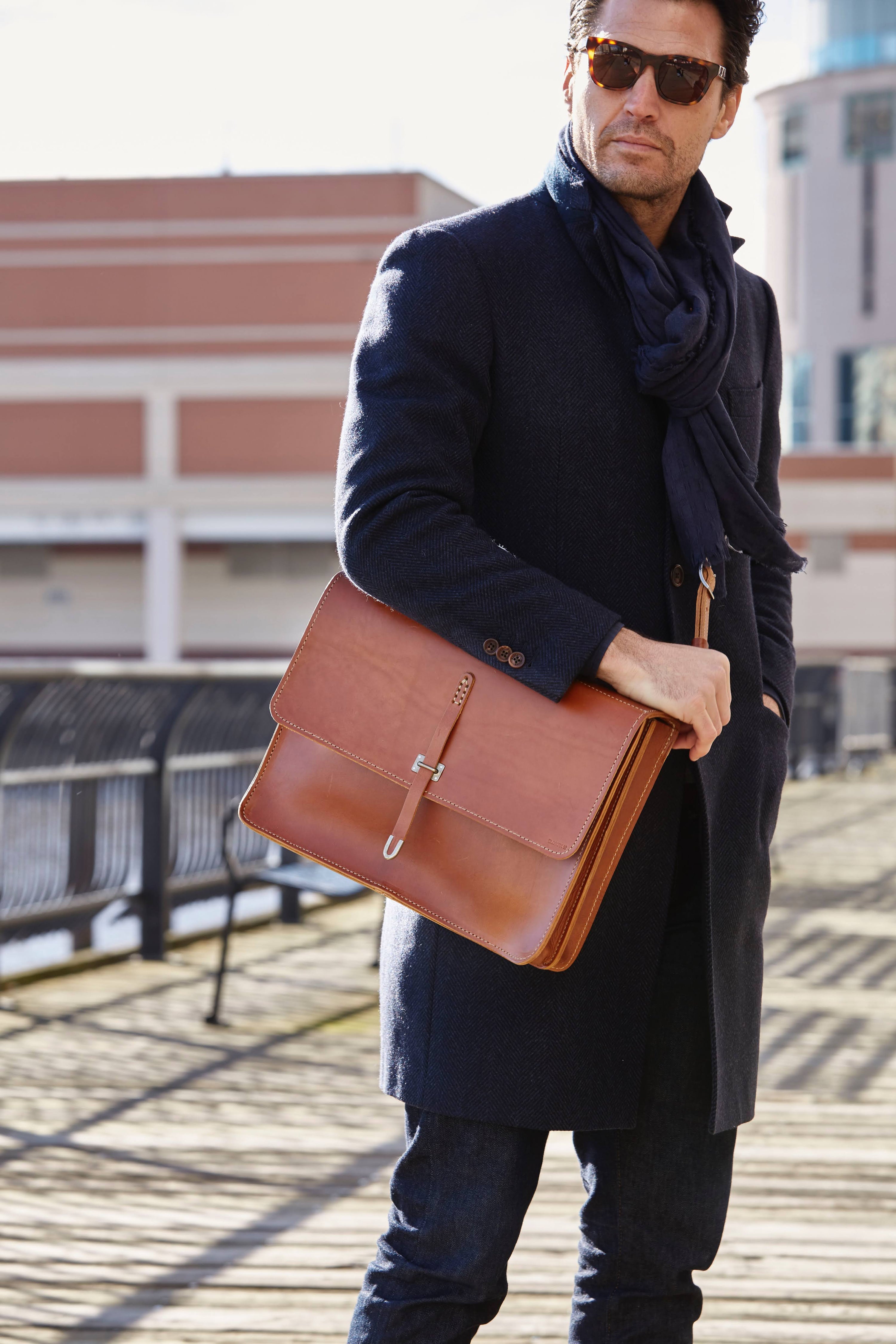 A man wearing sunglasses, a dark coat, and scarf stands outdoors holding a large brown leather briefcase, with buildings and a railing in the background.