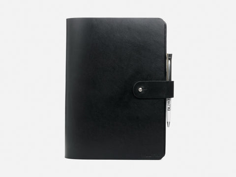 A closed black leather notebook with a snap button strap, featuring a silver pen clipped to the strap, set against a plain white background.
