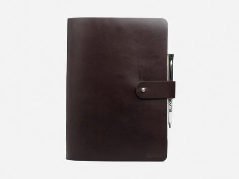 A closed, dark brown leather notebook with a snap button closure and a silver pen tucked into the side pen loop, set against a plain white background.