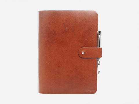 A closed brown leather notebook with a snap button closure and a black pen attached to the side, set against a plain white background.