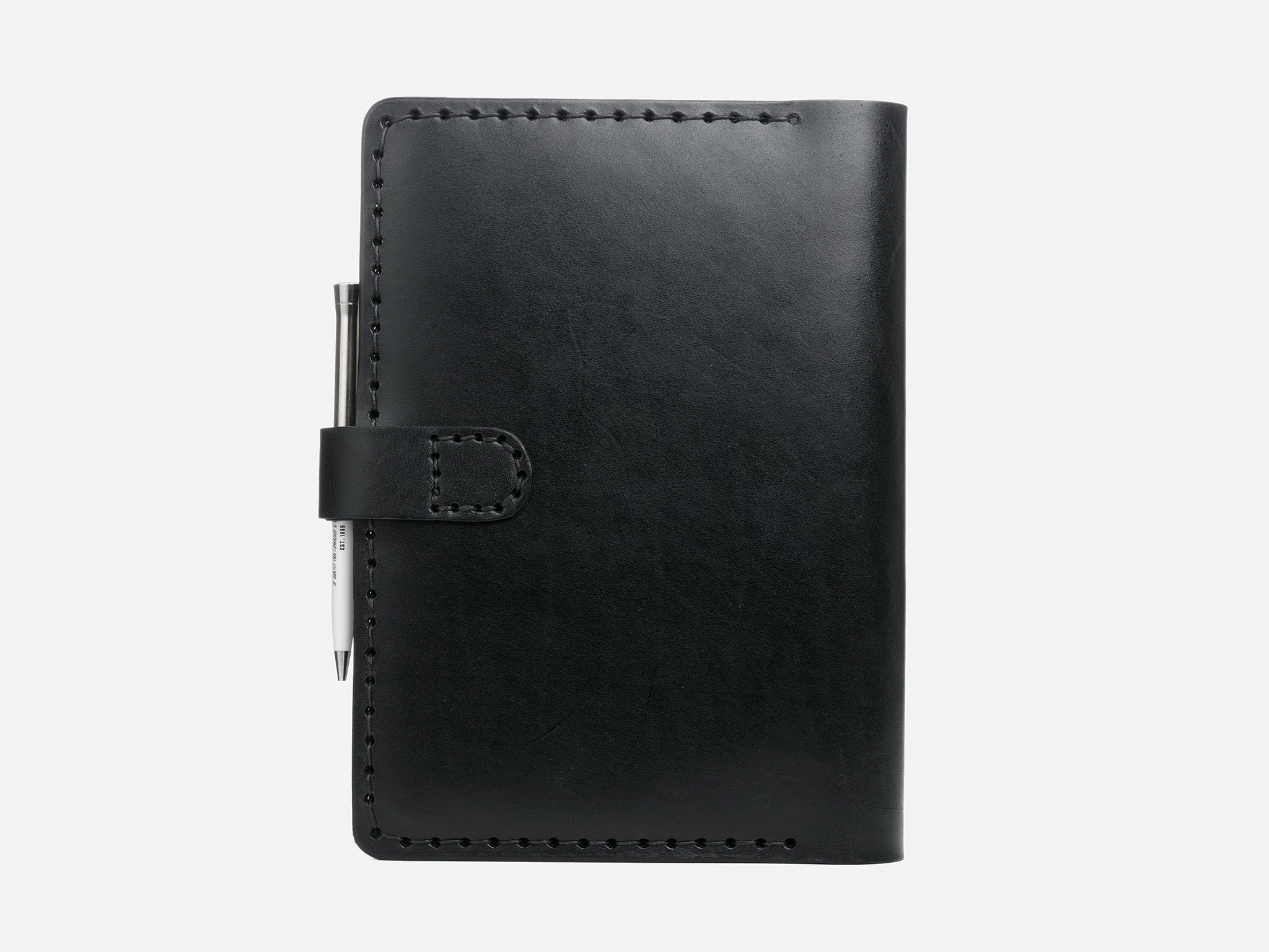 A closed black leather notebook with visible stitching along the edges and a metal pen secured under a strap on the left side, set against a plain white background.