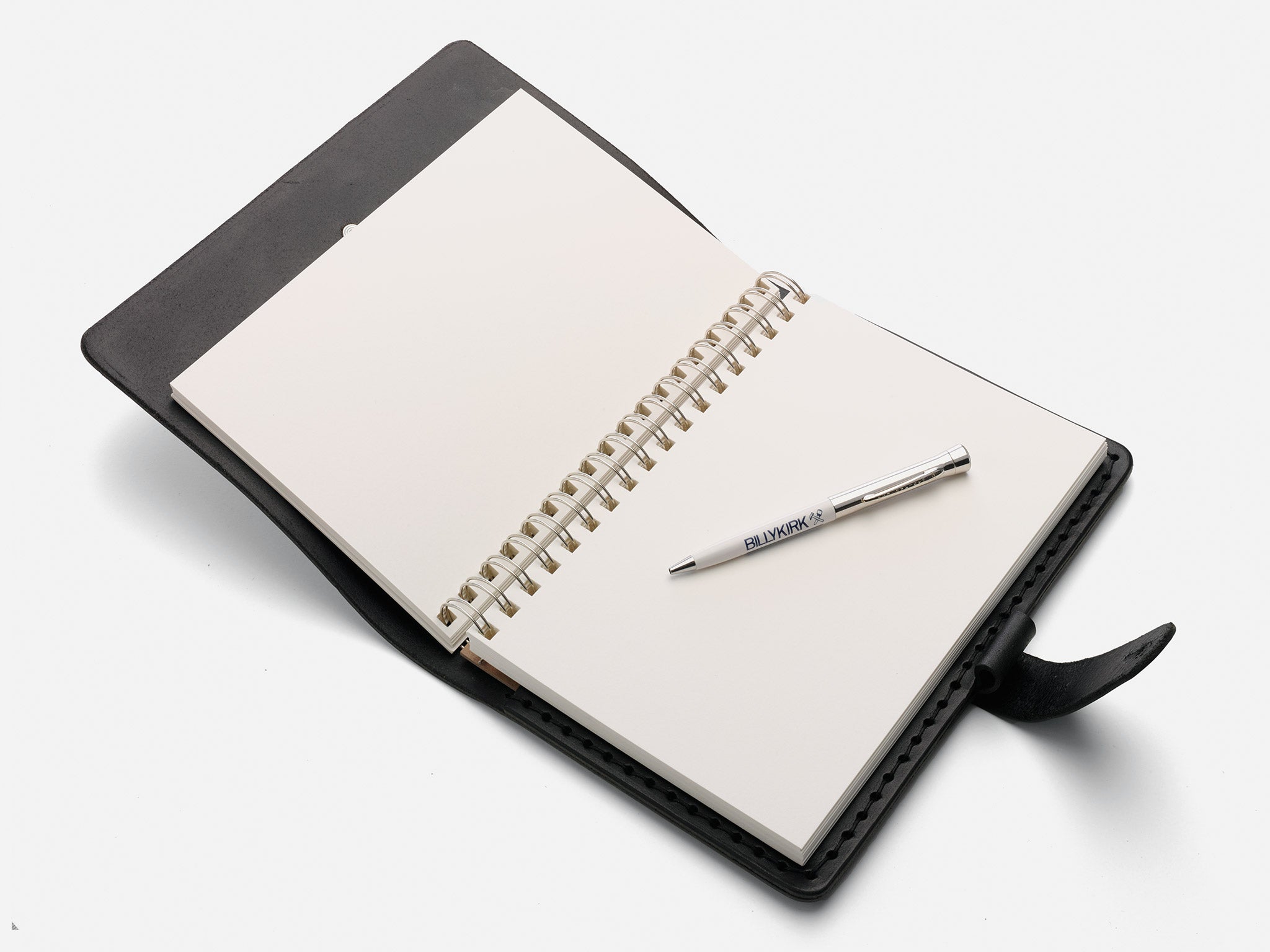 An open spiral-bound notebook in a black cover lies on a white surface, with a silver pen placed diagonally across the blank pages.